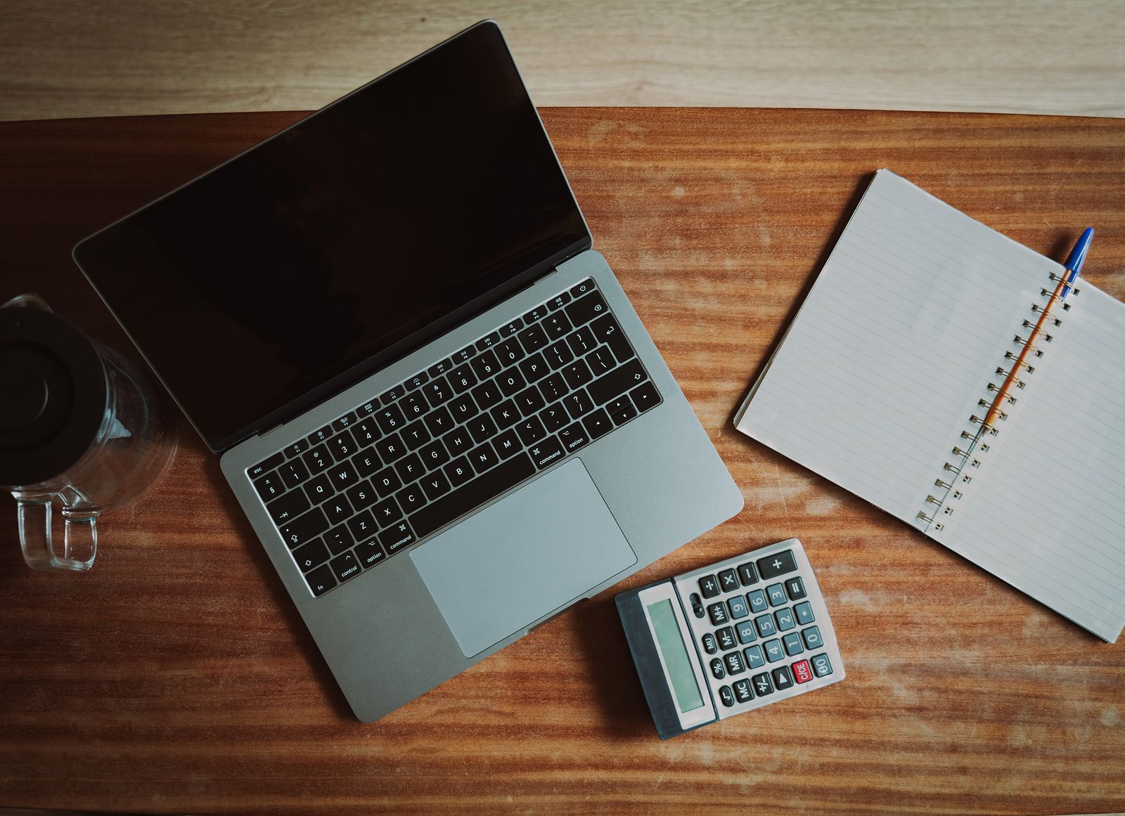 A laptop, calculator and open notebook on a wooden desk, ready to plan weekly bookkeeping hours