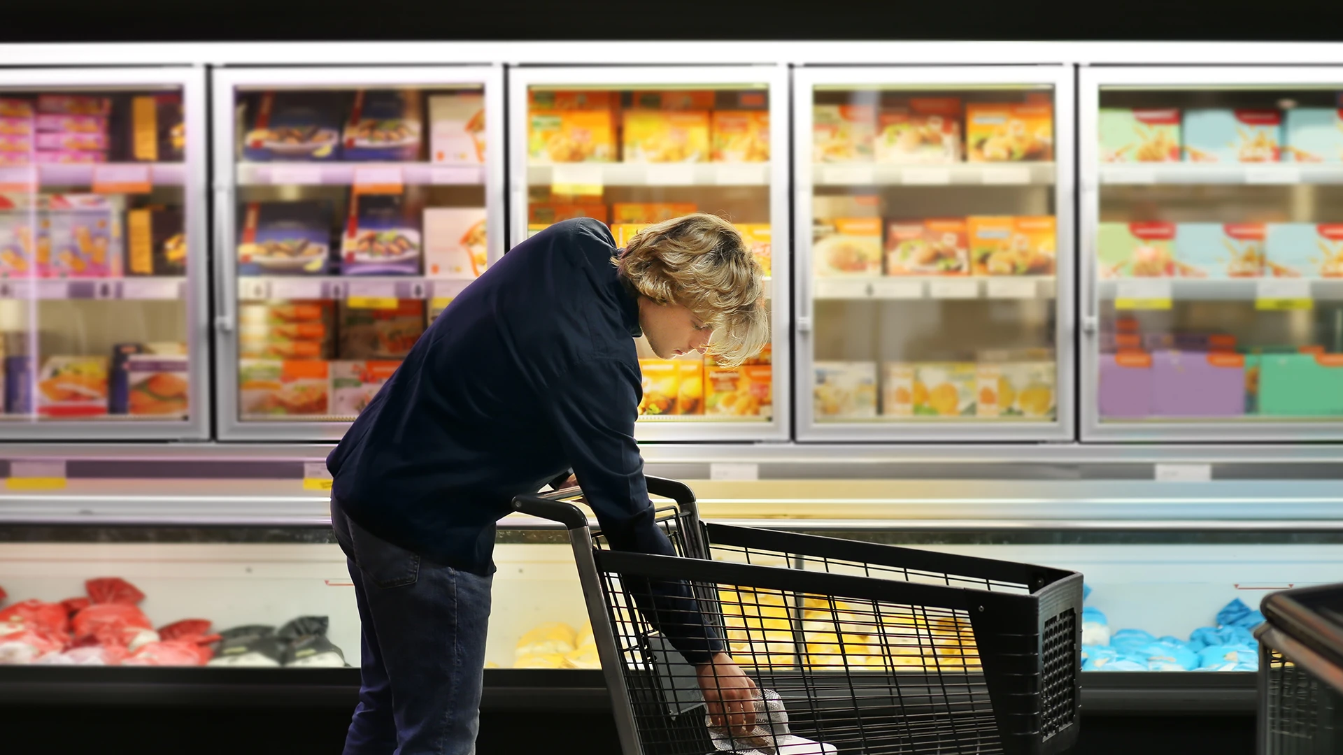 A man in his 30s reaches into a shopping card in the frozen food section