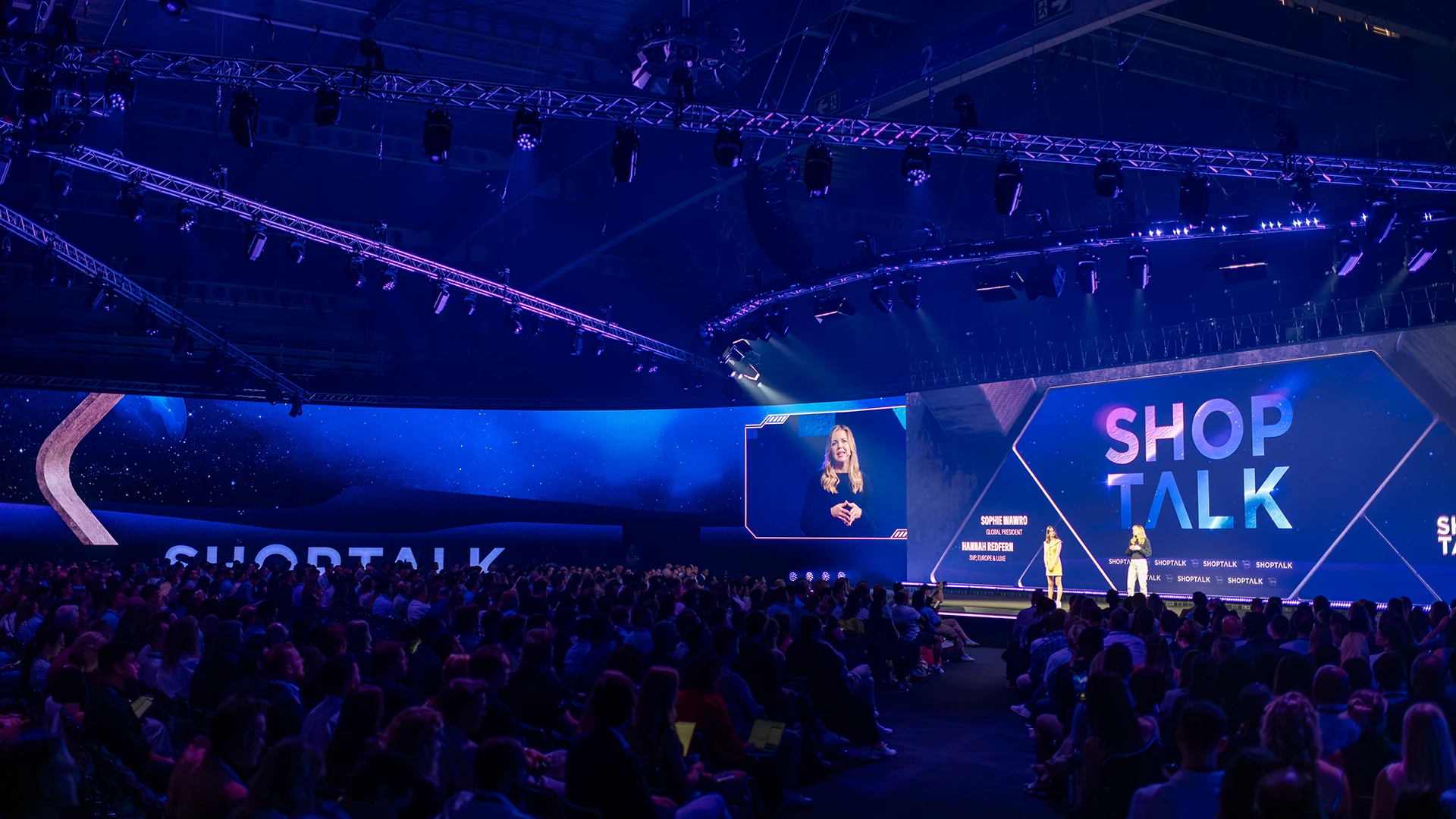 Large conference audience seated in a darkened arena facing a brightly lit stage with giant digital screens displaying “SHOP TALK.”