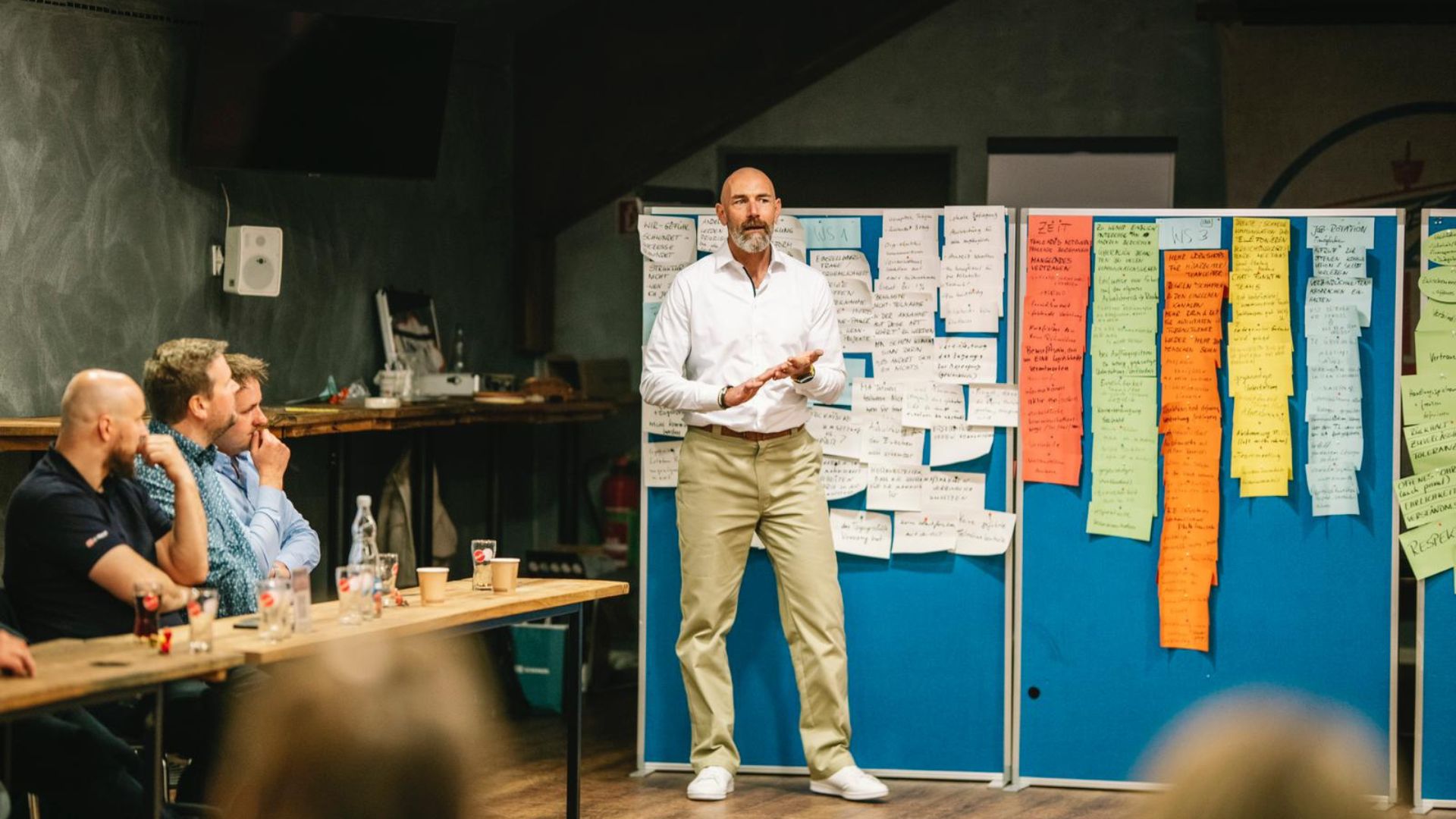 Mann hält Vortrag vor Pinnwand mit Notizzetteln, Teilnehmer hören zu.