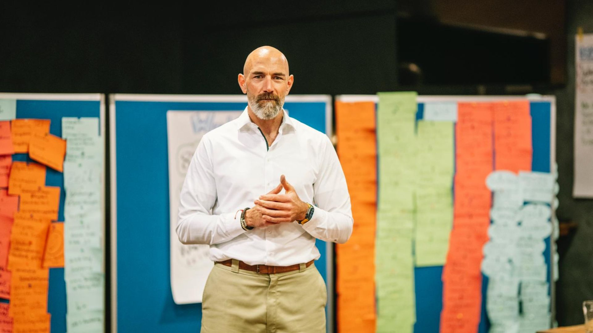 Mann in weißem Hemd steht vor farbenfroher Pinnwand und spricht konzentriert während eines Workshops