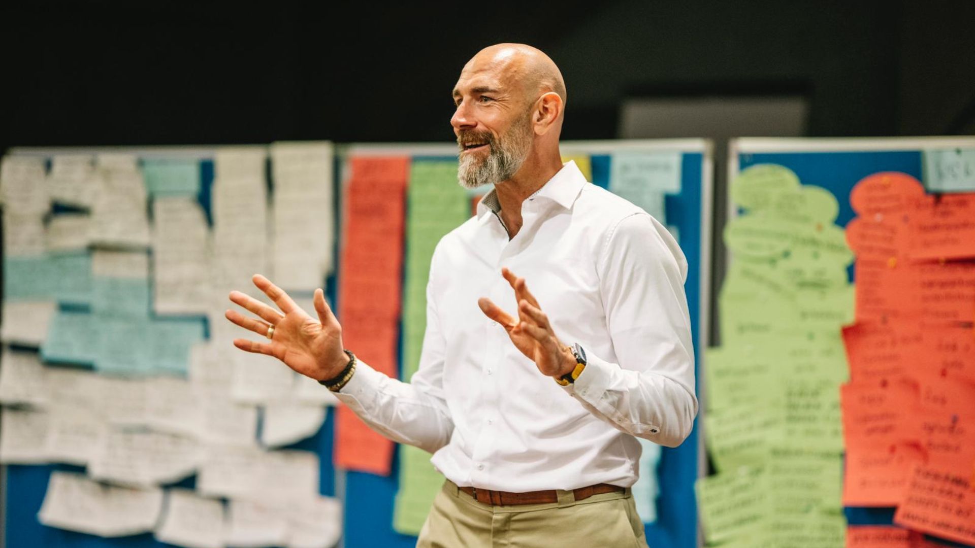 Lächelnder Mann in weißem Hemd spricht gestikulierend vor einer Pinnwand voller bunter Notizzettel.