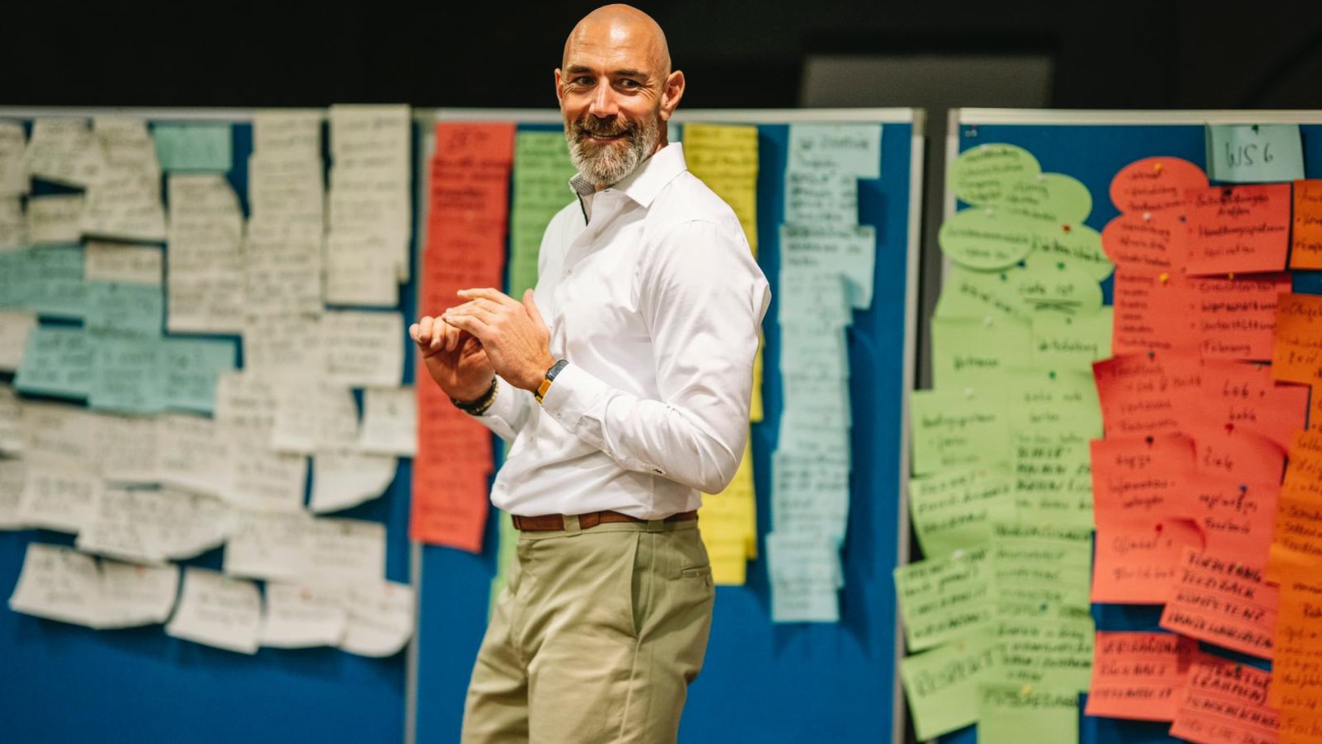 Vertriebstrainer schaut lächelnd zur Gruppe, während er vor einer bunt bestückten Moderationswand mit vielen Notizzetteln steht.