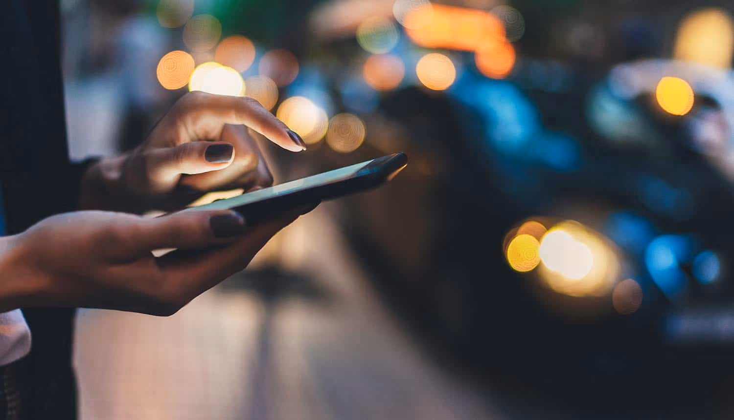 Mains de femme tenant un smartphone, ongles vernis bordeaux, lumières floues de rue nocturne