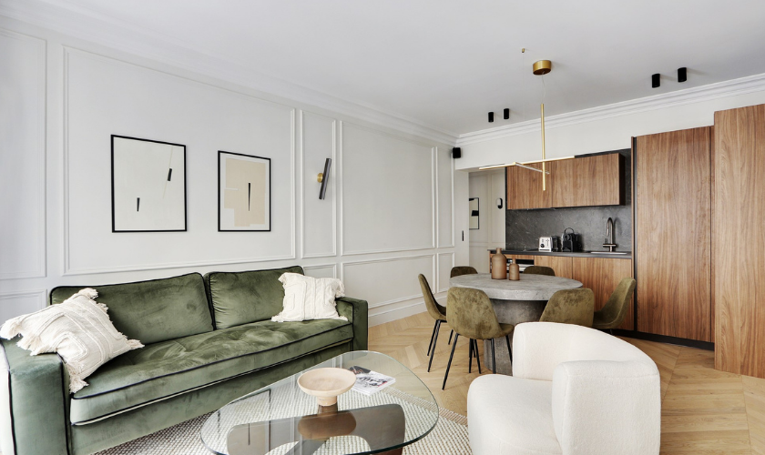 Salon moderne avec canapé vert, table basse en verre et cuisine ouverte en bois dans un appartement lumineux