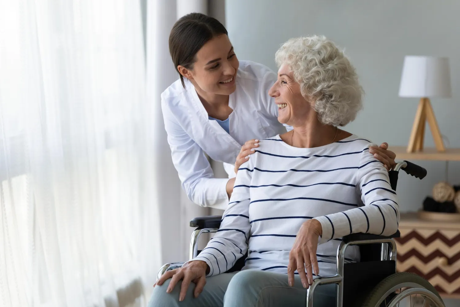 Smiling female caregiver comforting elderly woman in wheelchair by gently placing hands on her shoulders.