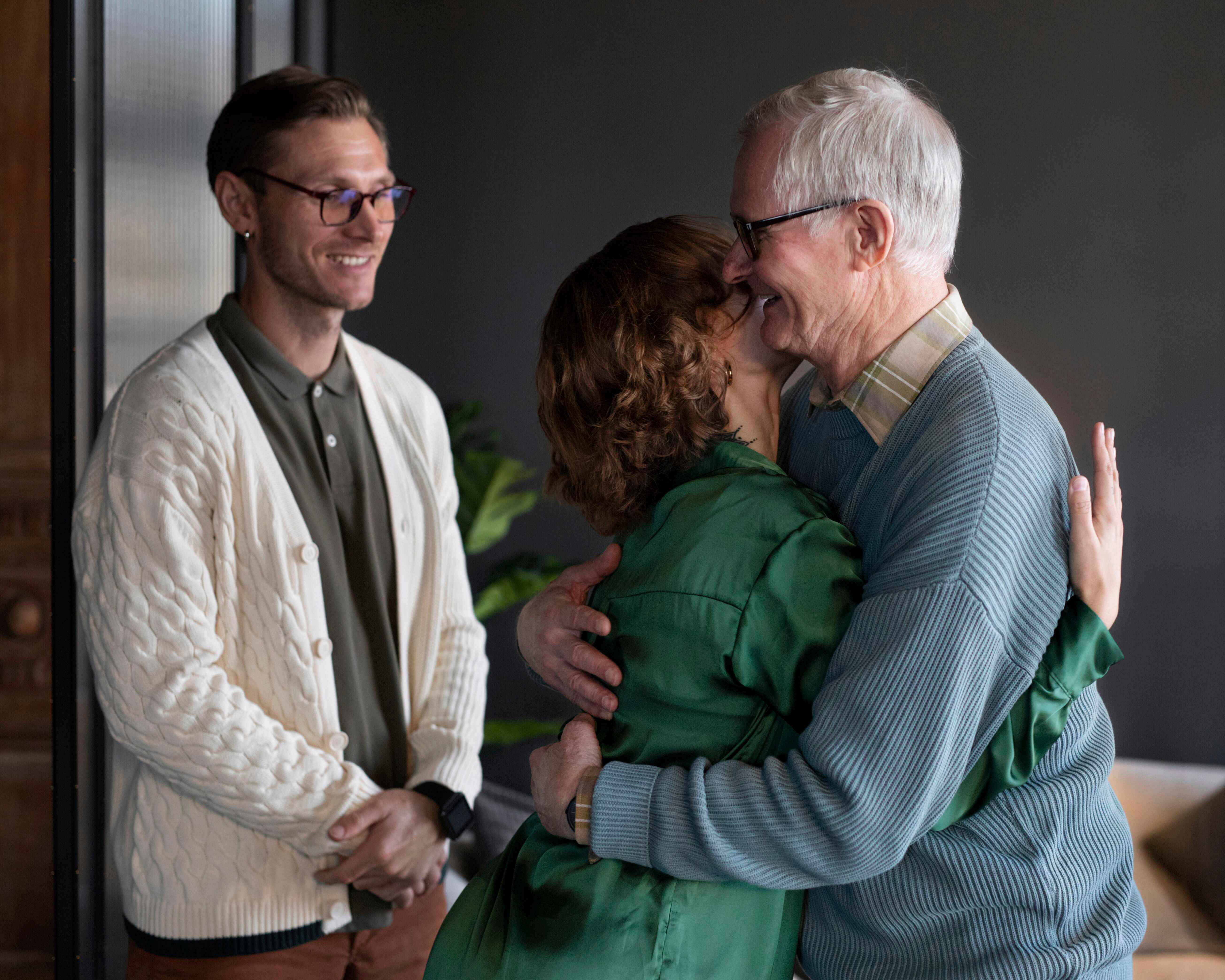 An elderly man in glasses and a blue sweater hugs a woman in a green coat while a younger man in a white cardigan smiles nearby.