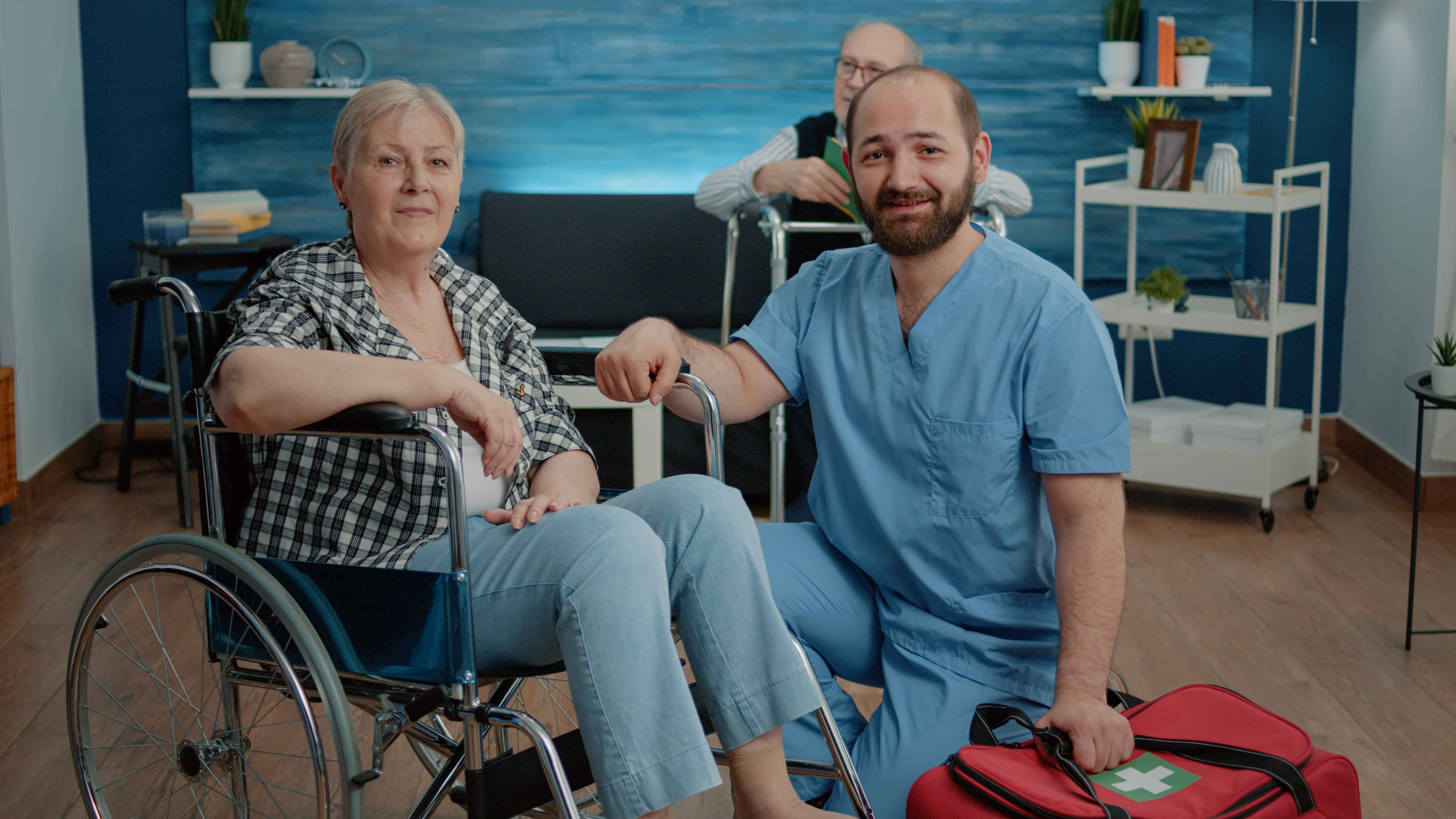 Elderly woman sitting in a wheelchair with a male healthcare worker kneeling beside her holding a red first aid bag.