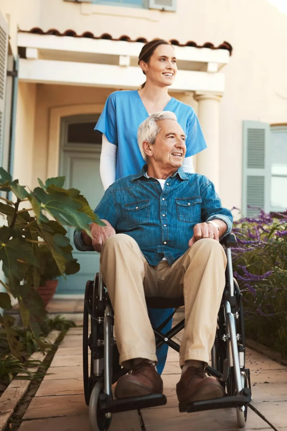 Smiling elderly man in a wheelchair being pushed by a female caregiver in blue scrubs outside a home.