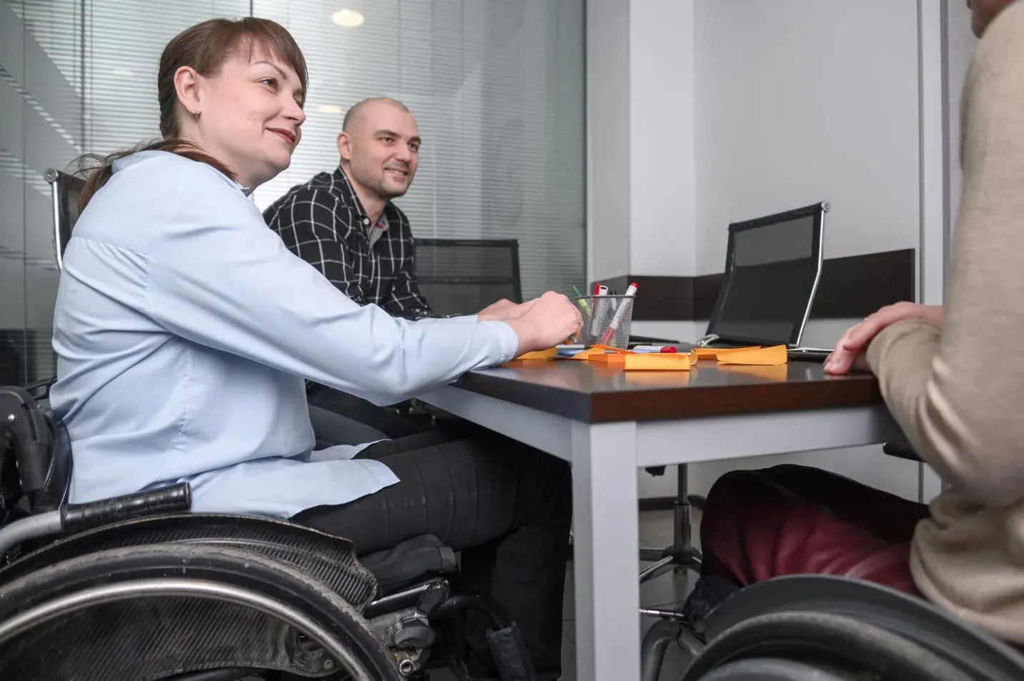 Three people in a meeting room, two in wheelchairs, sitting at a table with sticky notes and pens.