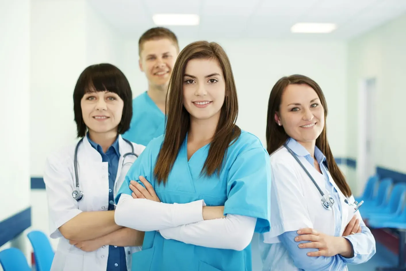 Group of four smiling medical professionals in scrubs and lab coats standing in a hospital corridor.