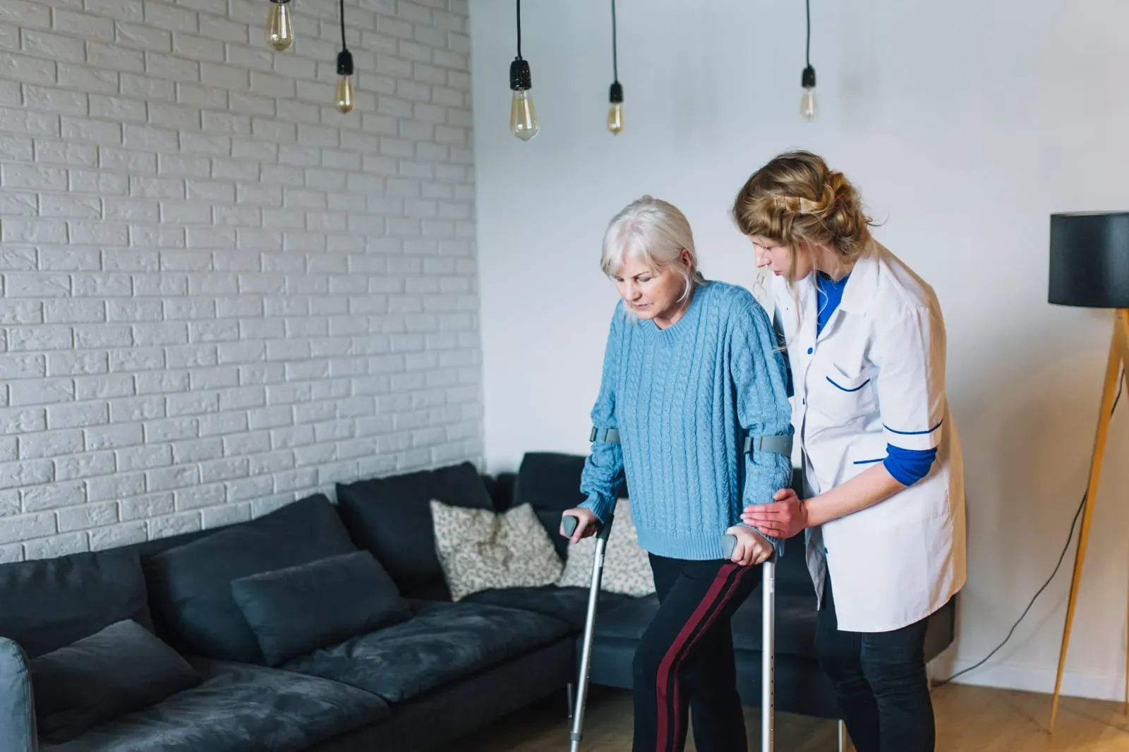 Young female caregiver assisting elderly woman with crutches in a living room.
