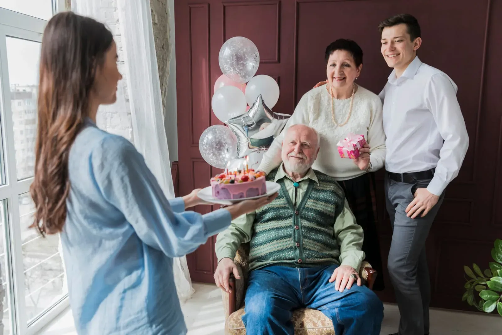 Woman holding a birthday cake with lit candles approaching an elderly man seated in a chair, accompanied by a smiling elderly woman holding a gift and a young man standing beside them with balloons in the background.