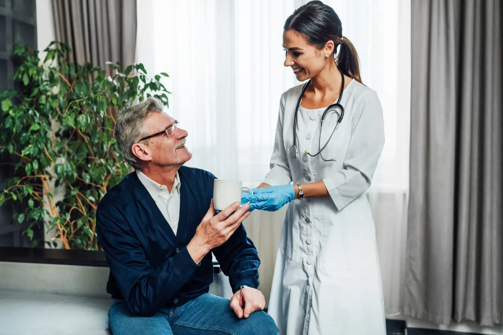 Smiling female healthcare worker in white coat and gloves handing a white mug to an elderly man sitting indoors.