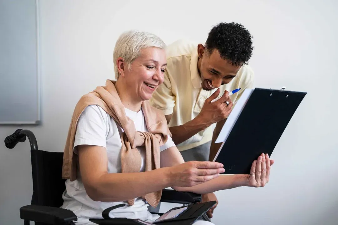 Smiling woman in wheelchair holding a clipboard with documents while a man stands beside her, both looking at the papers.