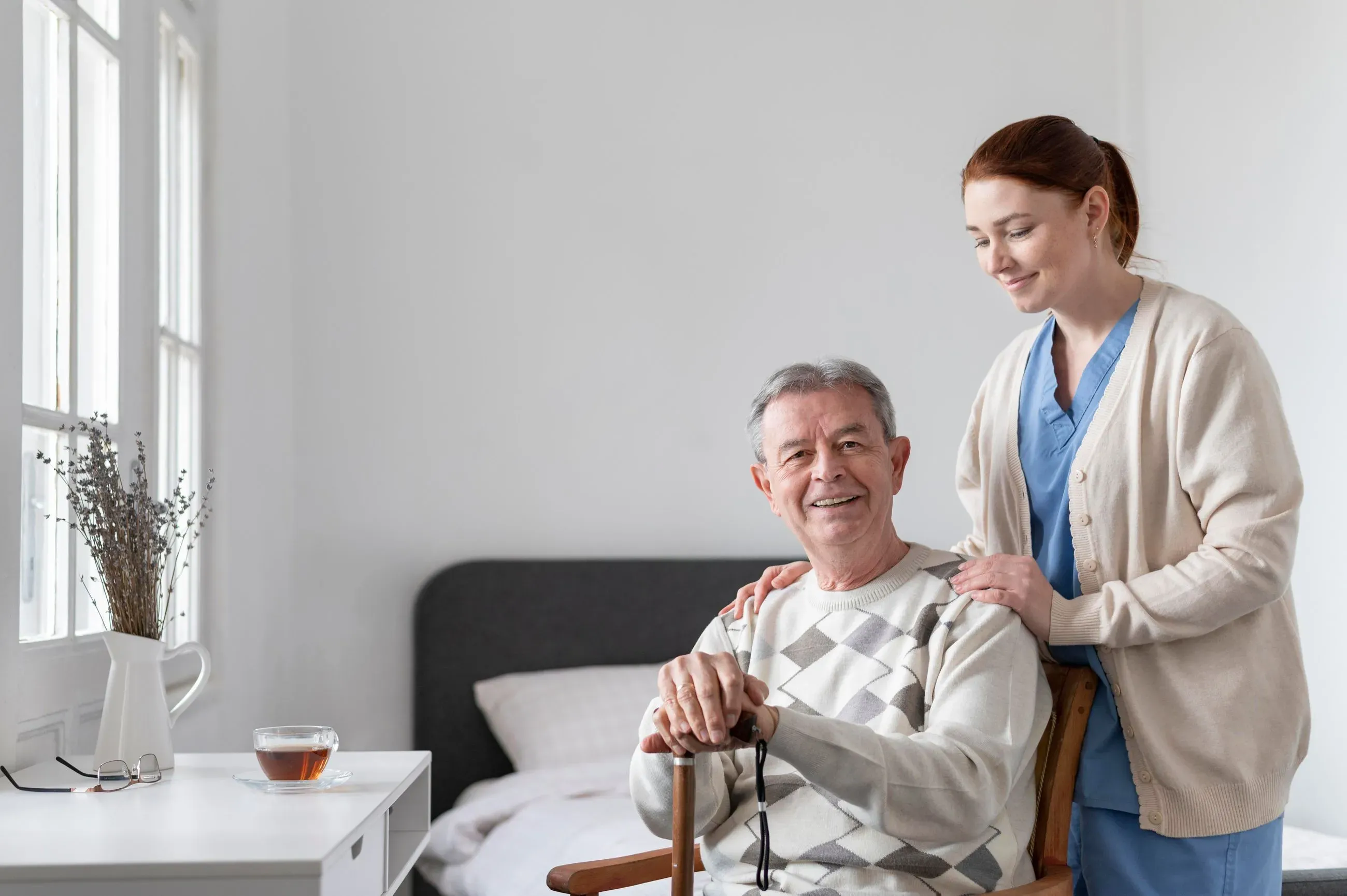 Senior man with cane sitting in a chair smiling, with a woman caregiver standing behind him with hands on his shoulders.