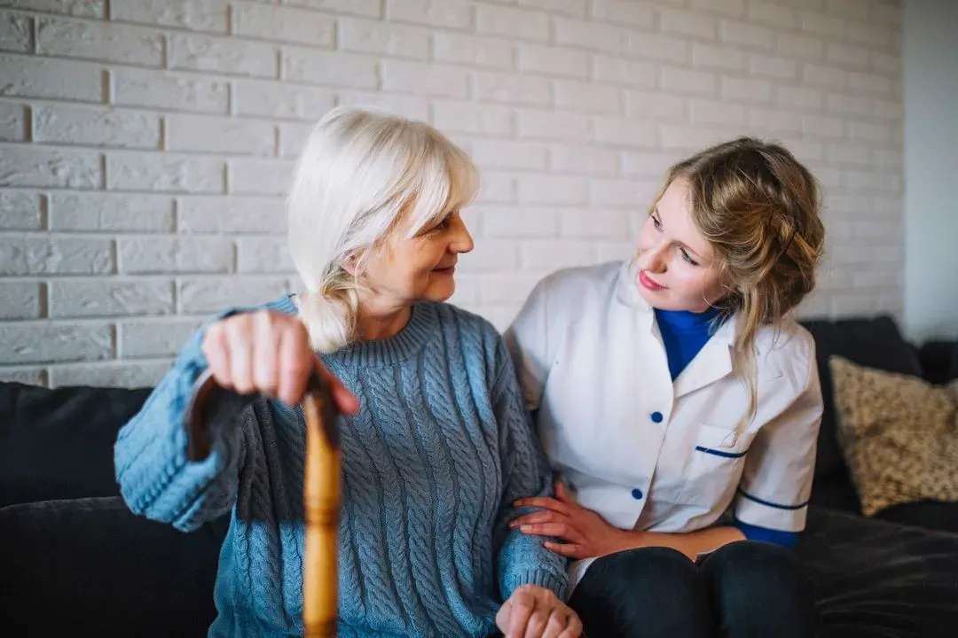 Young female caregiver sitting beside an elderly woman holding a wooden cane, both smiling and looking at each other.