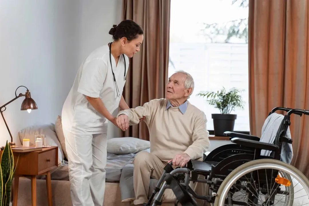 Caregiver assisting elderly man to stand up from bed near a wheelchair in a well-lit room.