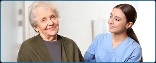 Smiling elderly woman with gray hair wearing a dark green shawl sitting next to a young female nurse in blue scrubs.