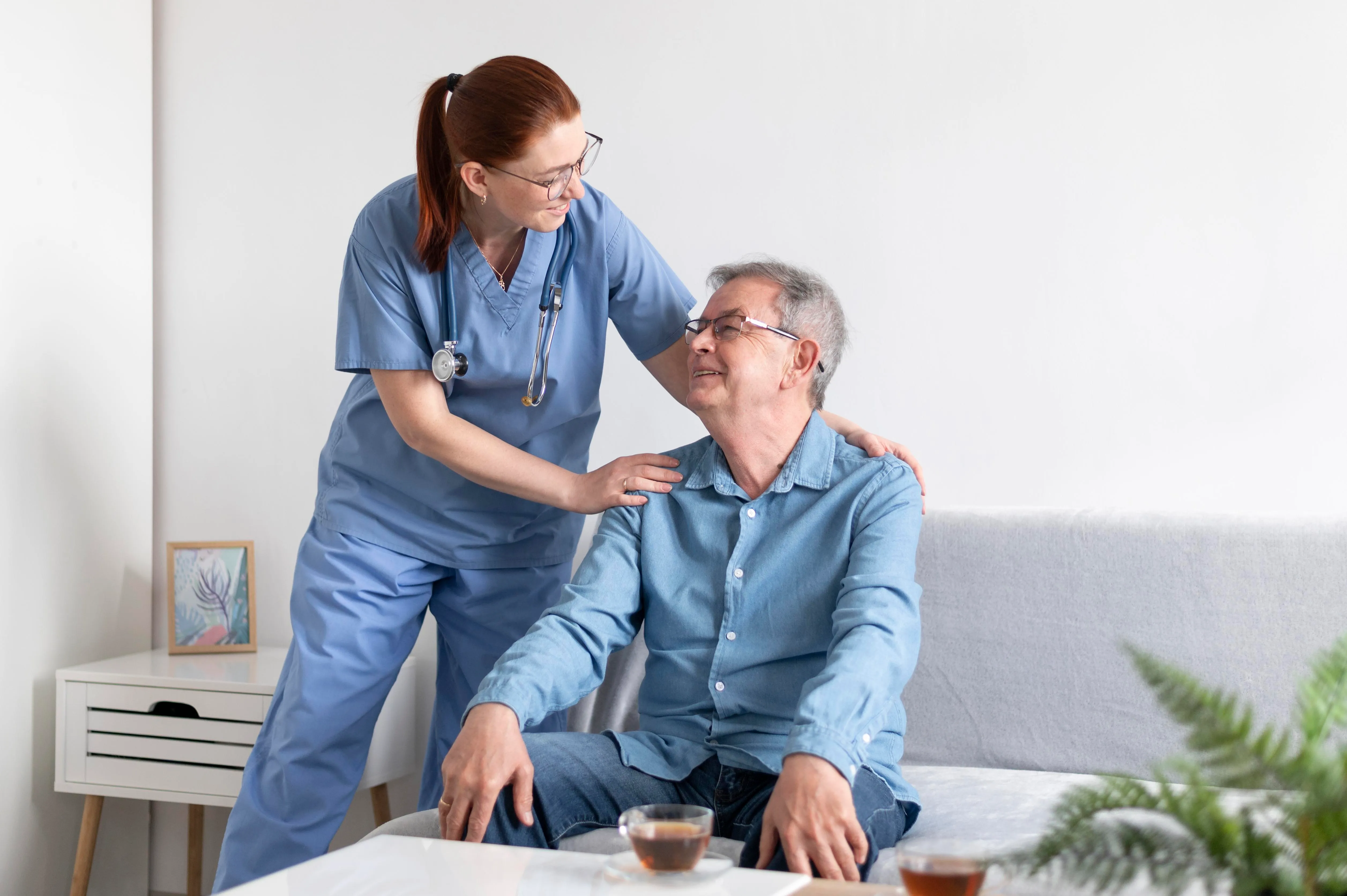 A nurse assists an elderly man sitting on a couch, providing support and care in a comfortable setting.