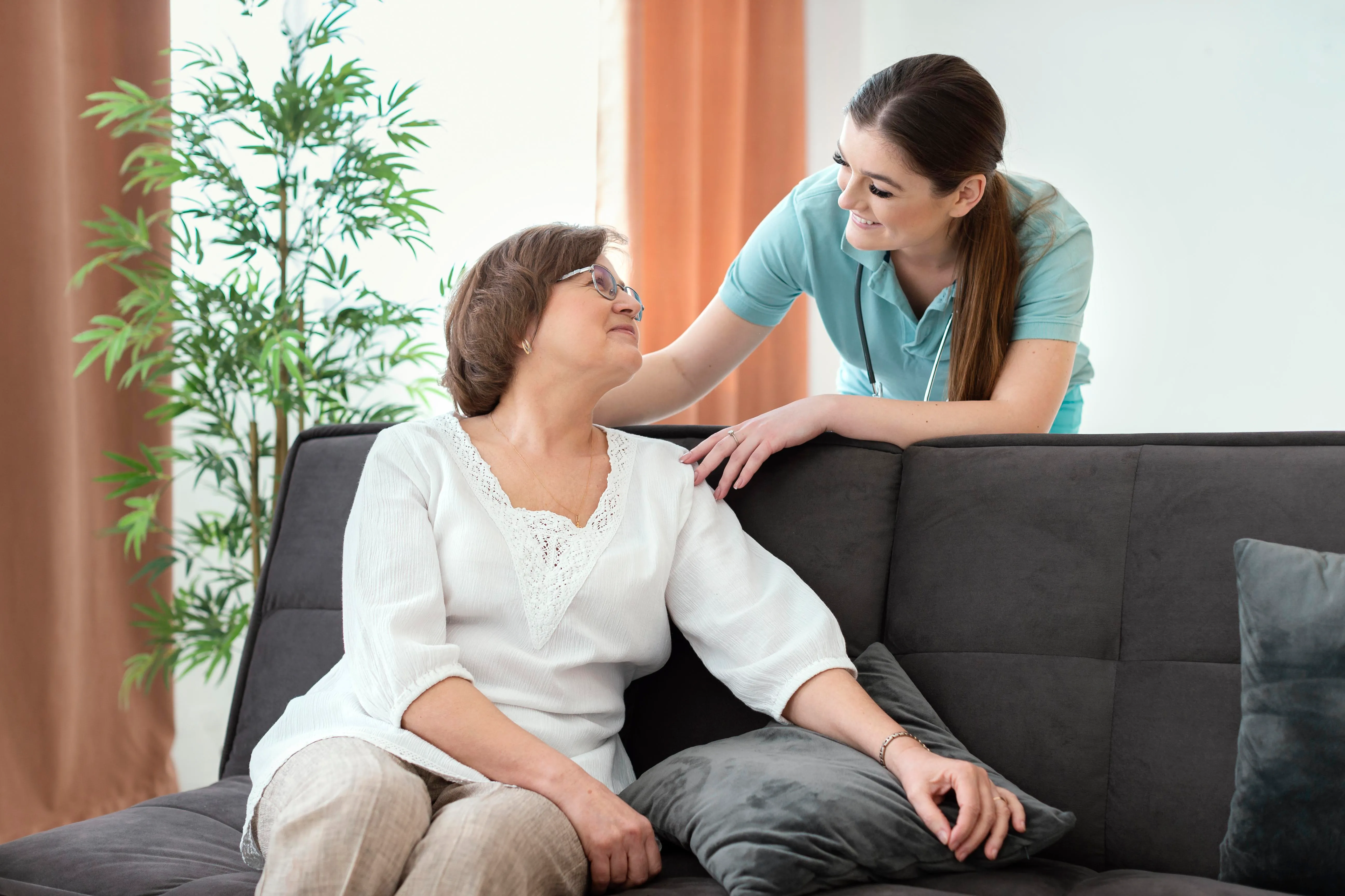 A woman sits on a couch with a caregiver, engaged in a supportive conversation in a cozy living room setting.