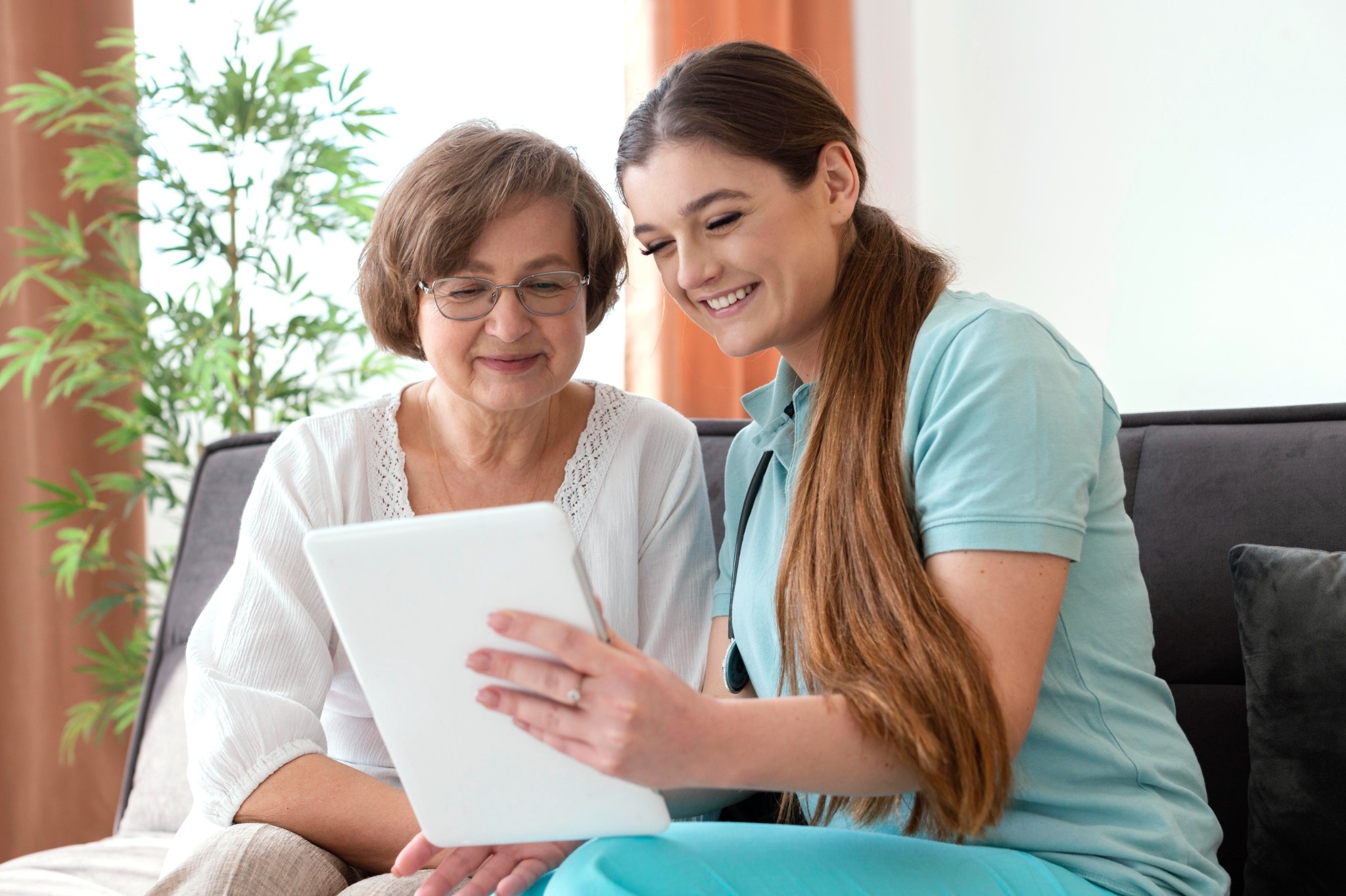 Smiling caregiver showing information on a tablet to an older woman while sitting together at home.