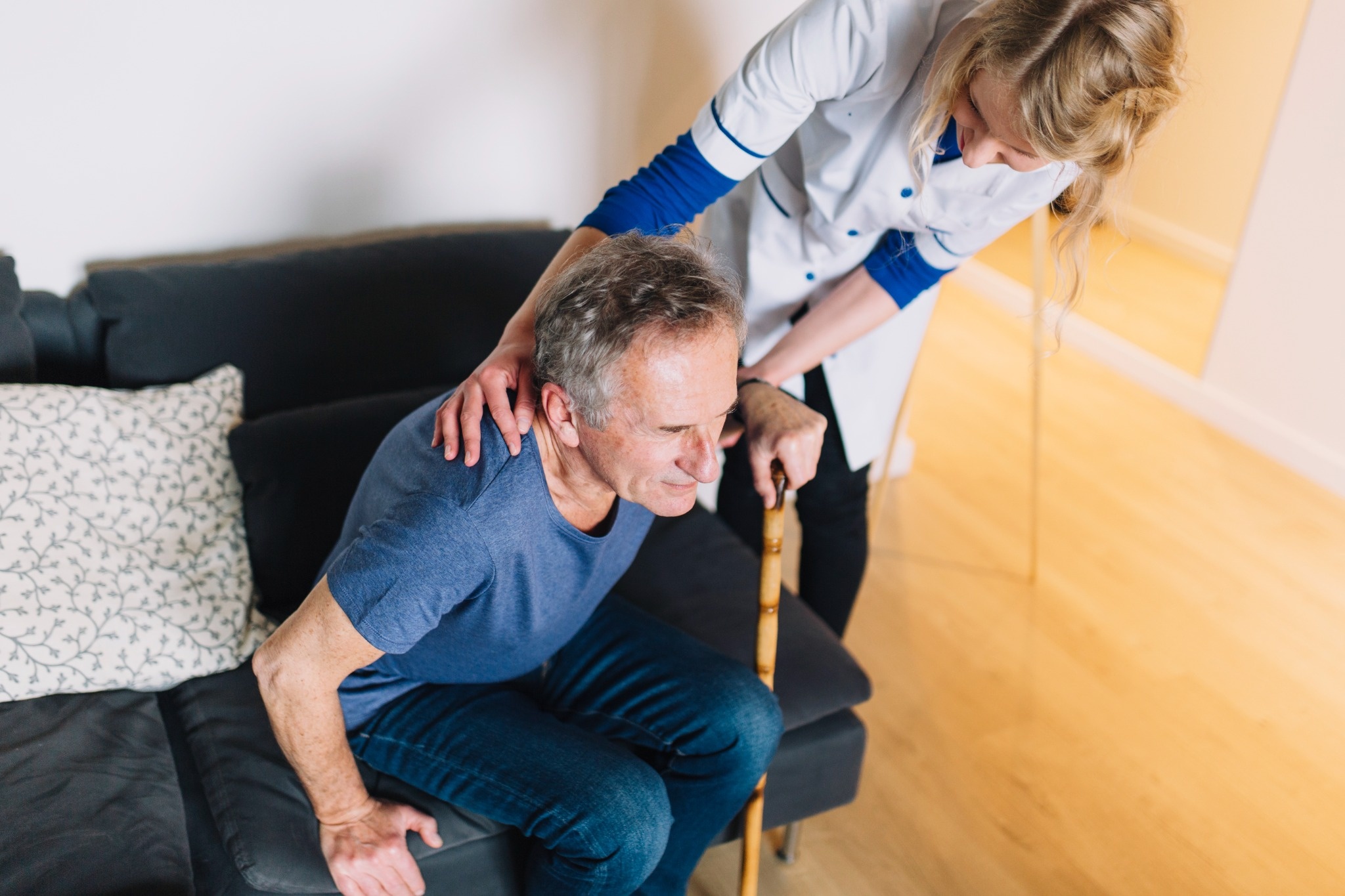Caregiver helping an elderly man stand up from a sofa while he uses a walking stick at home.