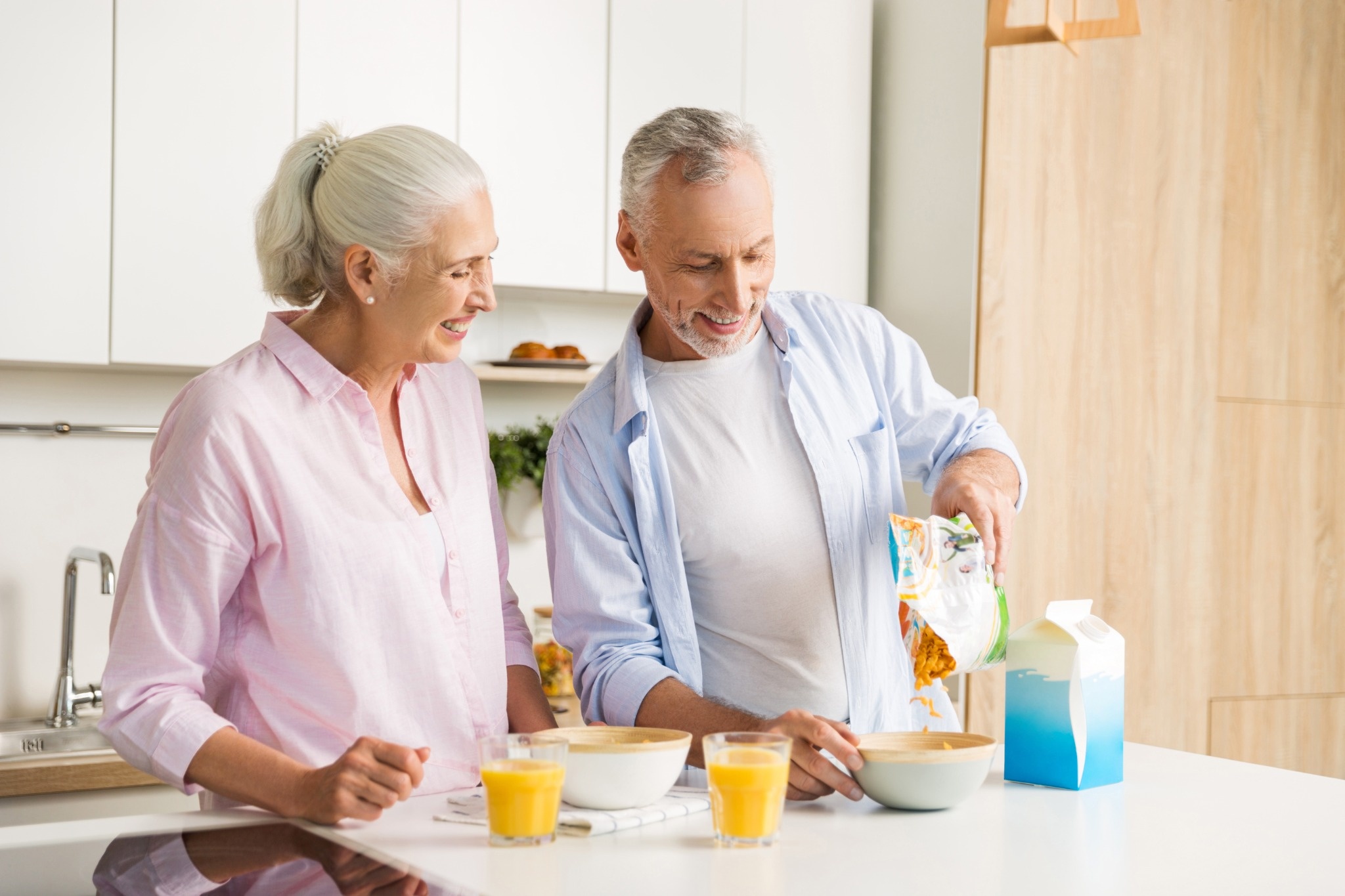 Smiling older couple preparing breakfast together in a bright kitchen with cereal and orange juice.