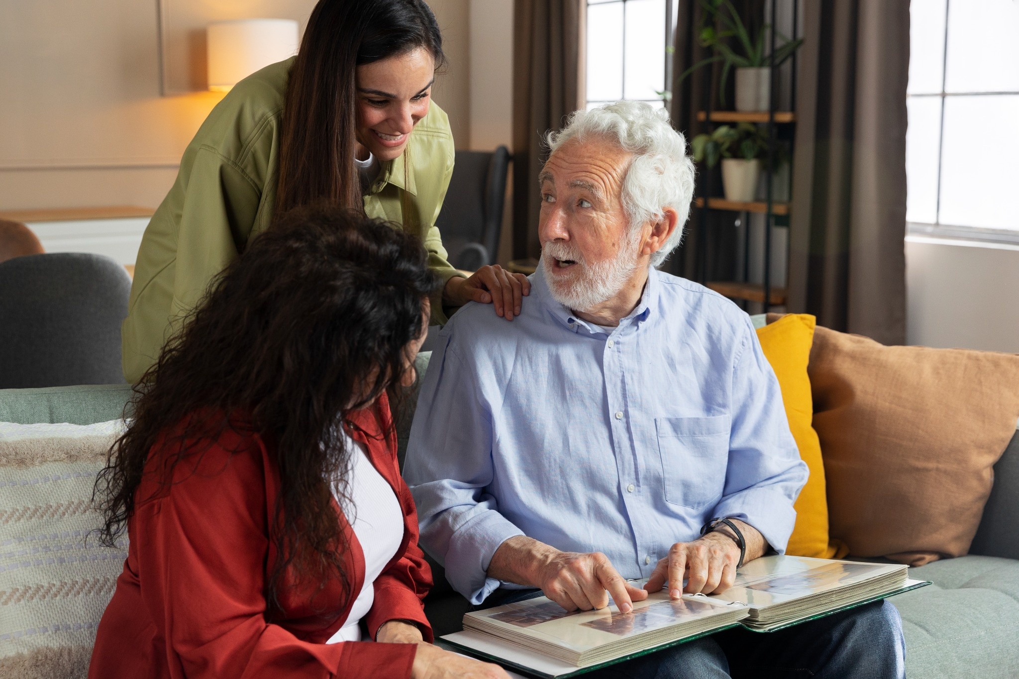 Elderly man looking through a photo album with two women while sitting together on a sofa at home.