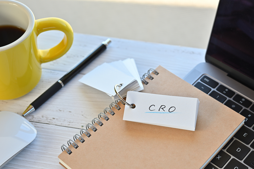Desk setup with a notepad labeled CRO (Conversion Rate Optimization), a pen, yellow coffee mug, and laptop — representing CRO strategy planning.