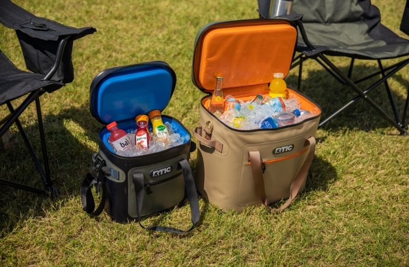 Two RTIC soft cooler bags filled with ice and drinks, open on a grassy outdoor setting with folding chairs in the background.