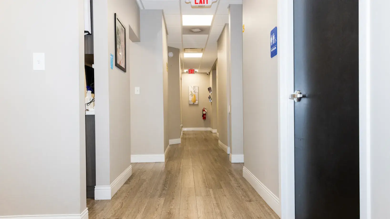 A long, empty hallway with wooden flooring and an exit sign above a door at the end.