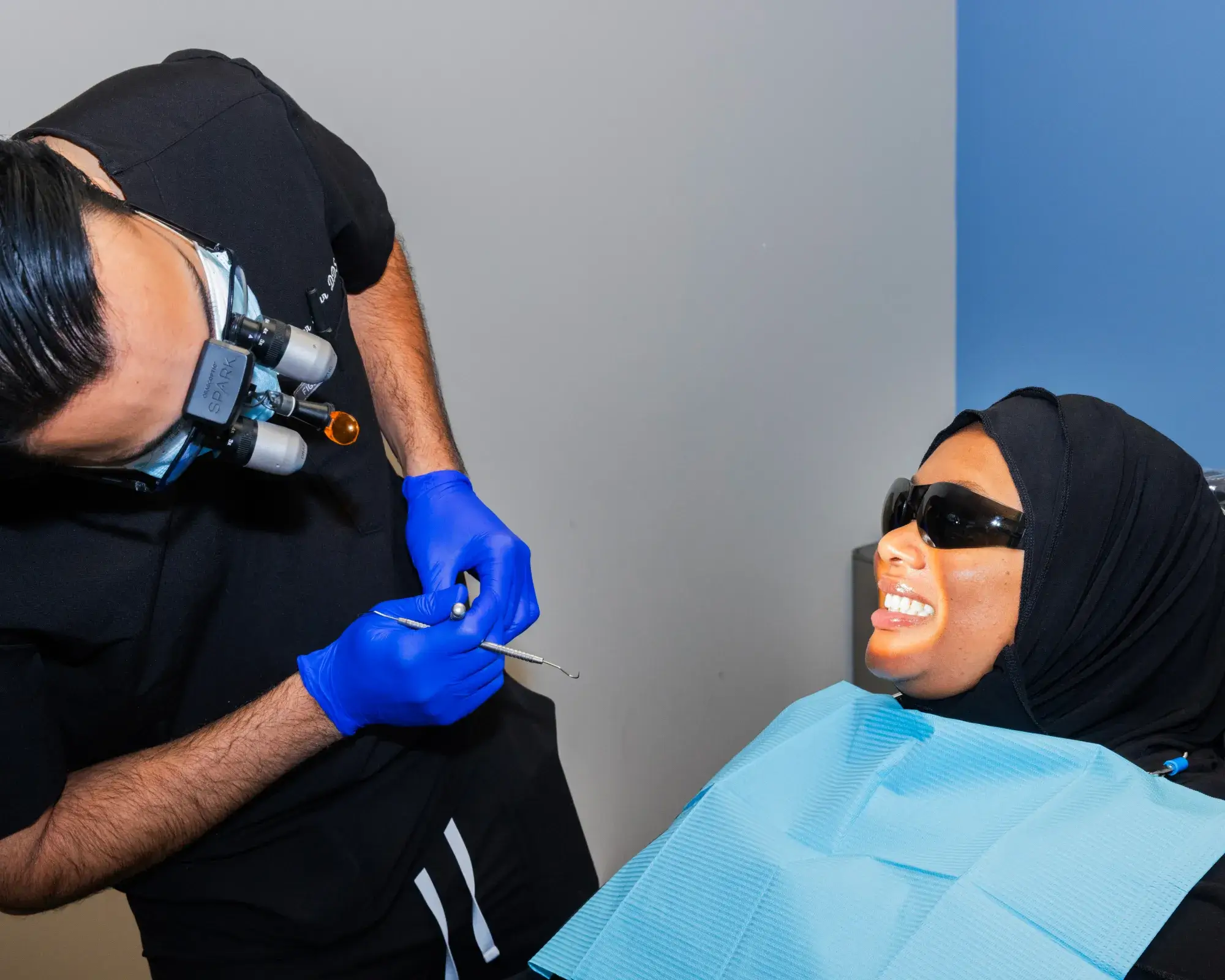 A dentist wearing magnifying glasses examines a patient with a dental tool as she smiles and wears sunglasses.
