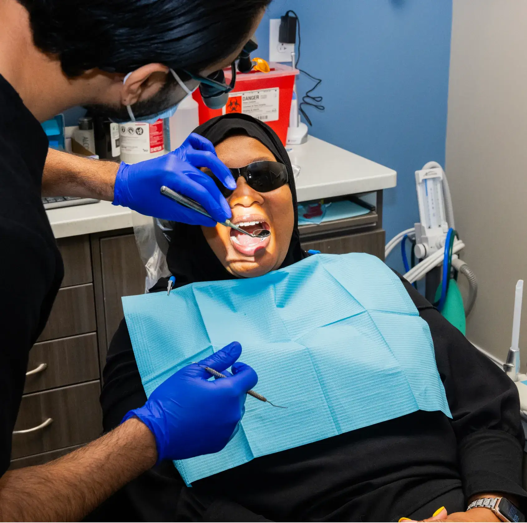 A dentist examines a patient's teeth using dental tools while she wears sunglasses and a bib.