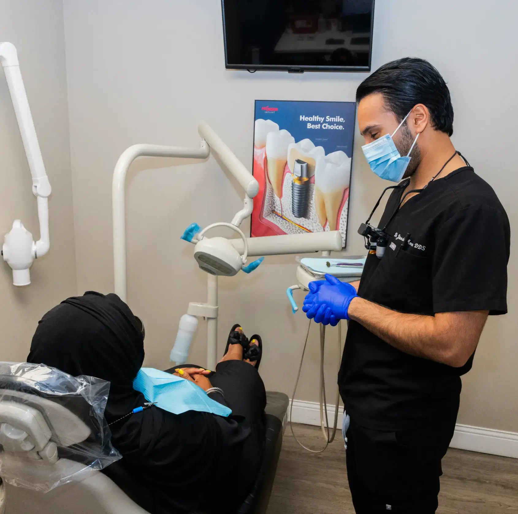 Dentist wearing mask and gloves talks to patient sitting in a dental chair.