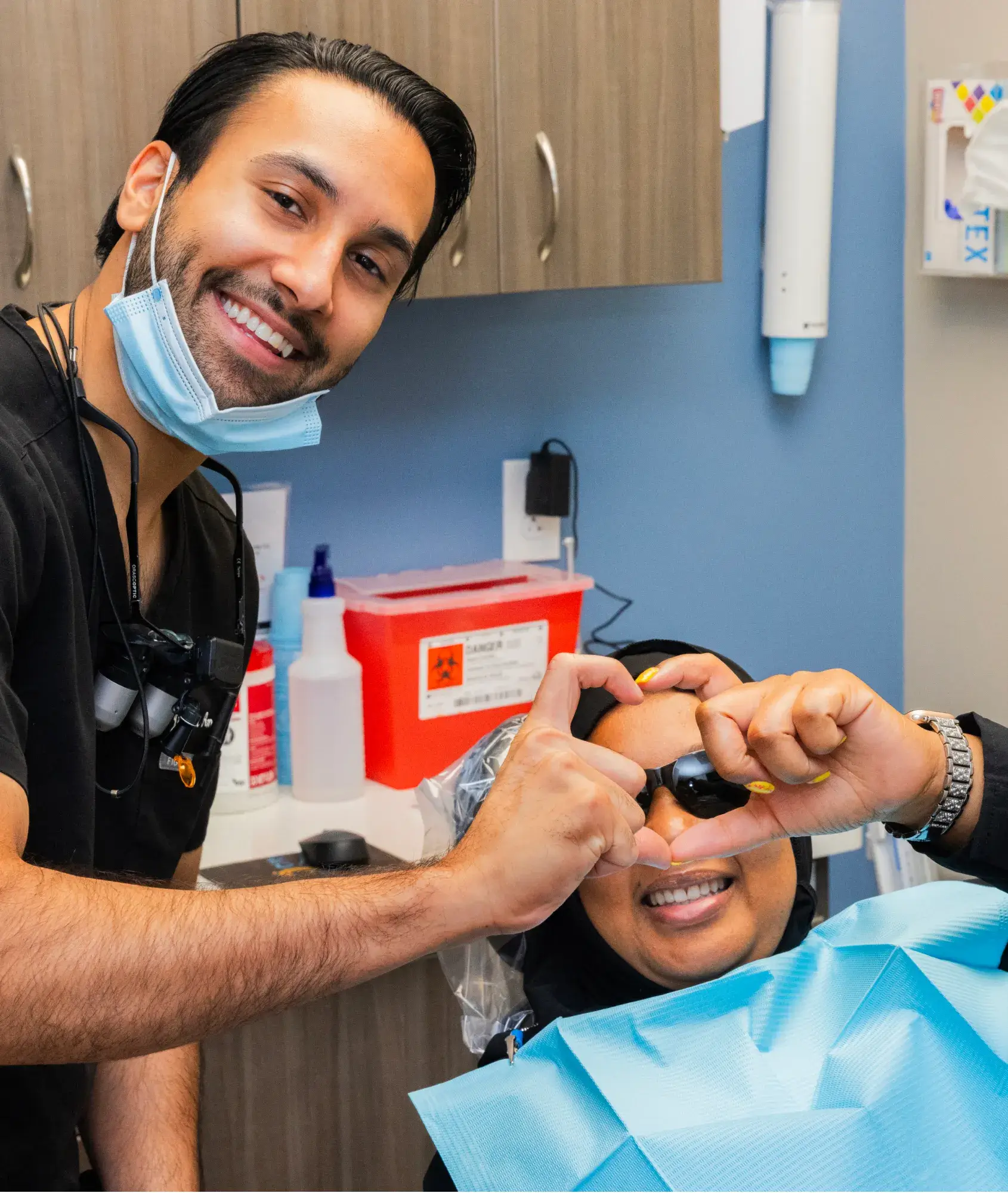 A dentist wearing a mask smiles, while a patient makes a heart shape with their hands.