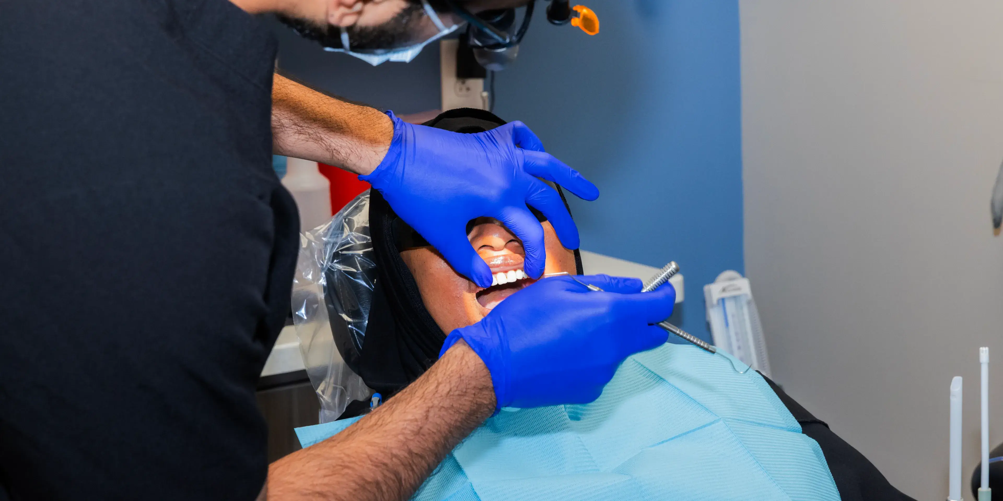Dentist wearing blue gloves examines woman's teeth in a dental chair.