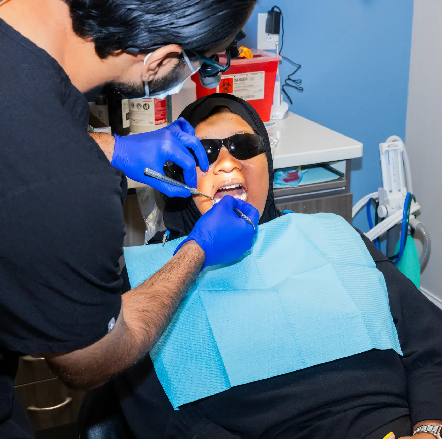 A dentist examines a patient's teeth, who wears sunglasses and a dental bib, in the clinic.