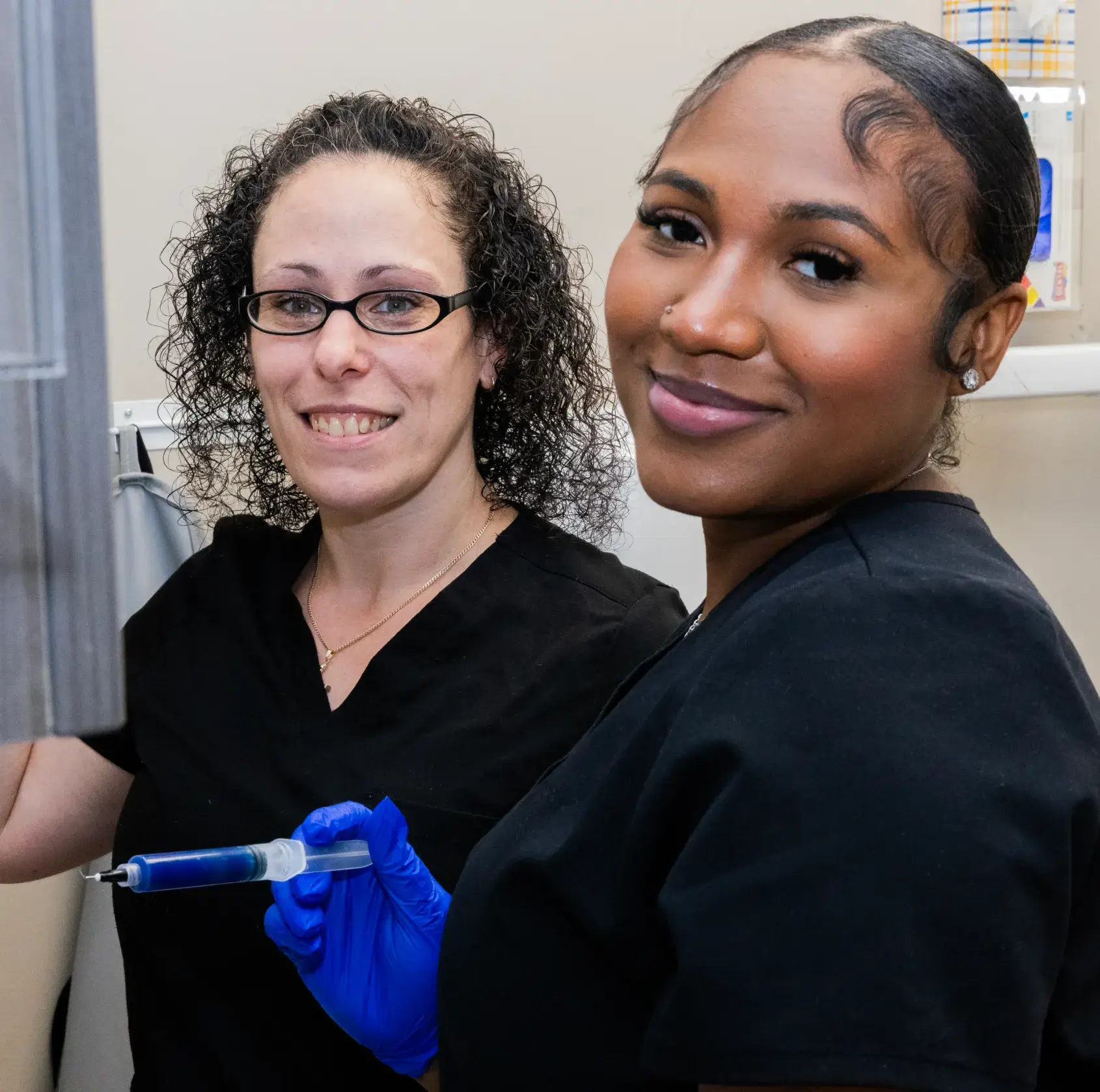 Two smiling healthcare workers in black scrubs, one holding a syringe, standing in a clinical setting.