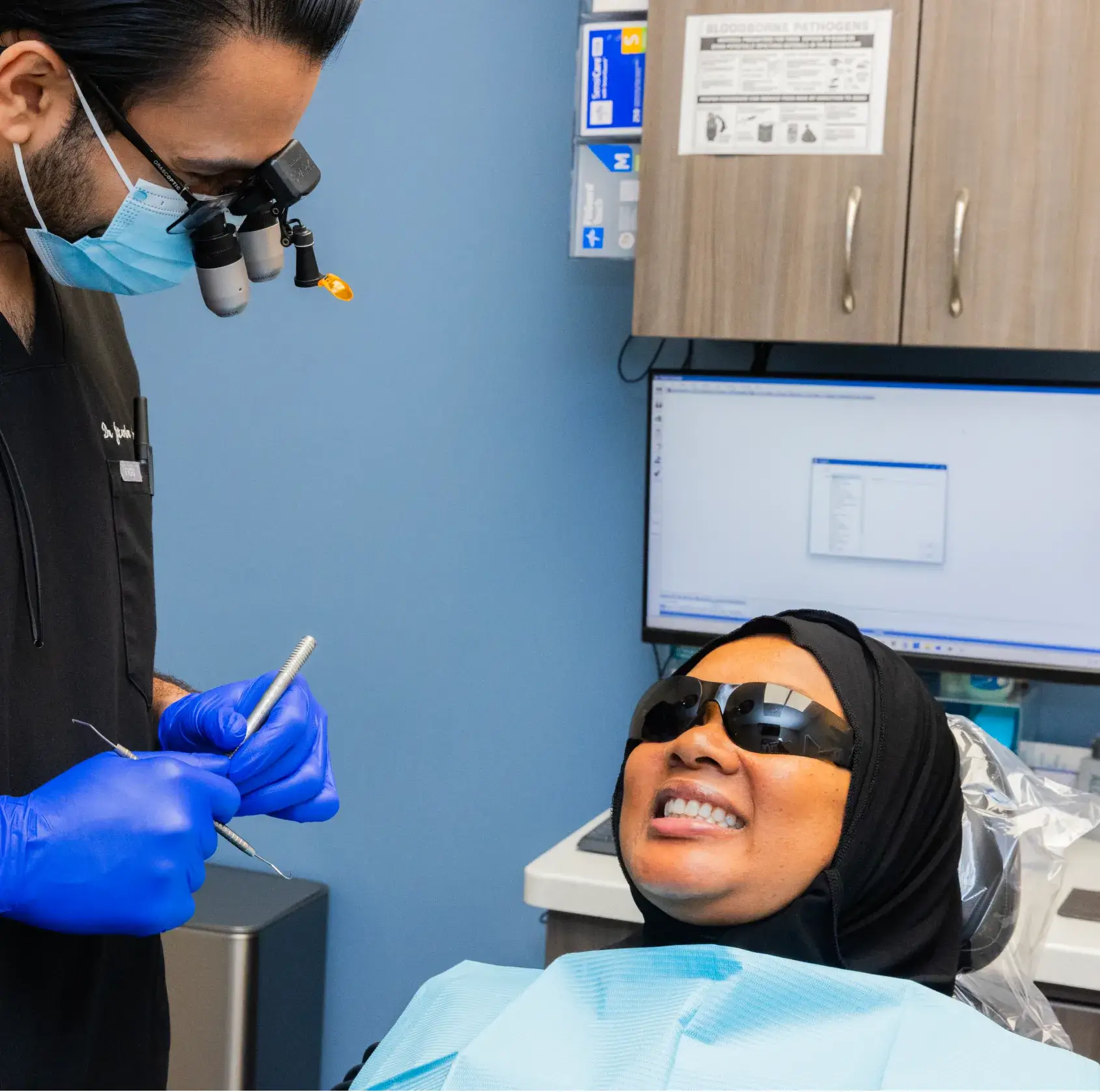 Dentist wearing magnifying glasses examines a smiling patient in a dental office.