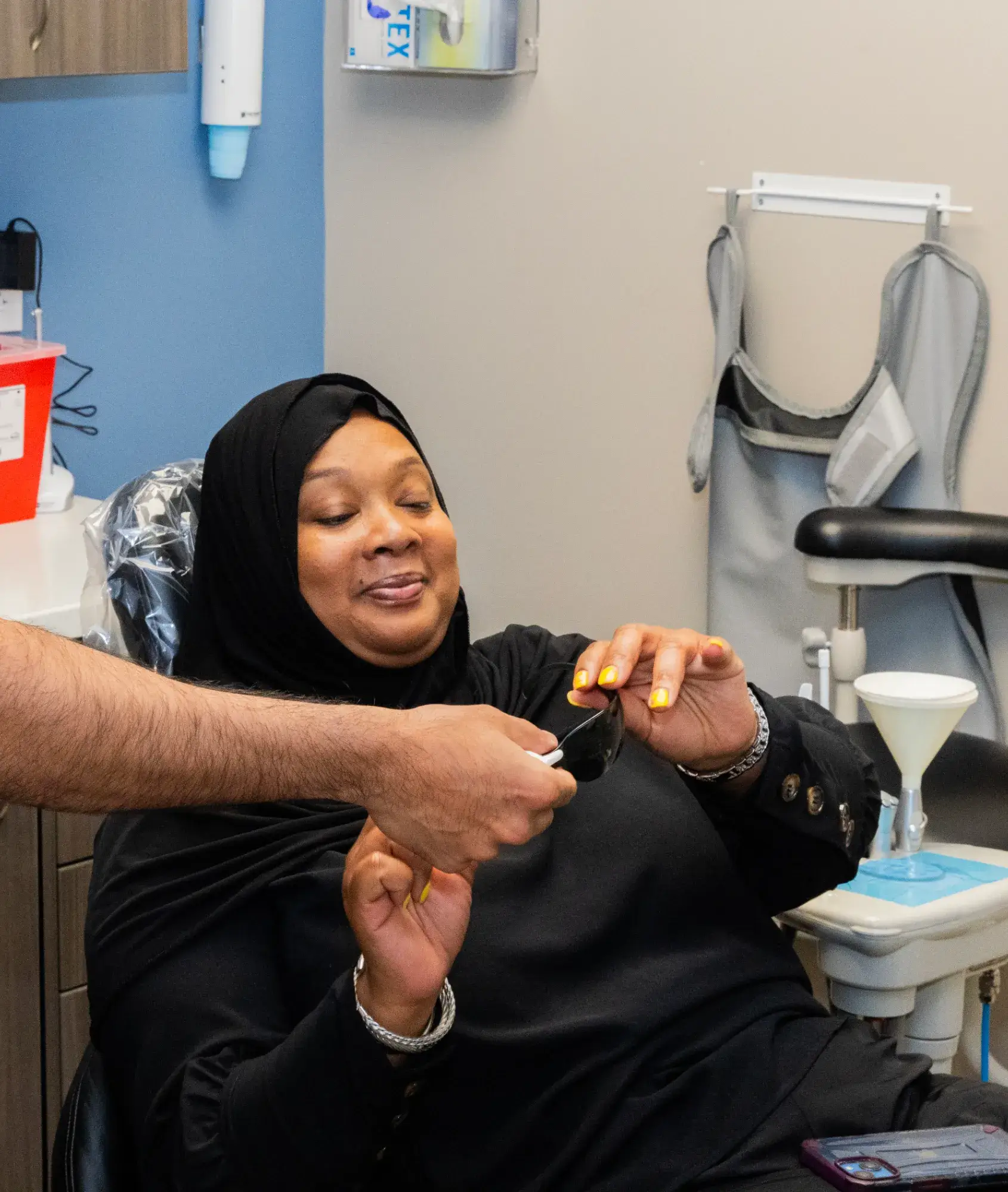 A woman in a dental chair smiles and holds dark glasses, assisted by a person next to her.