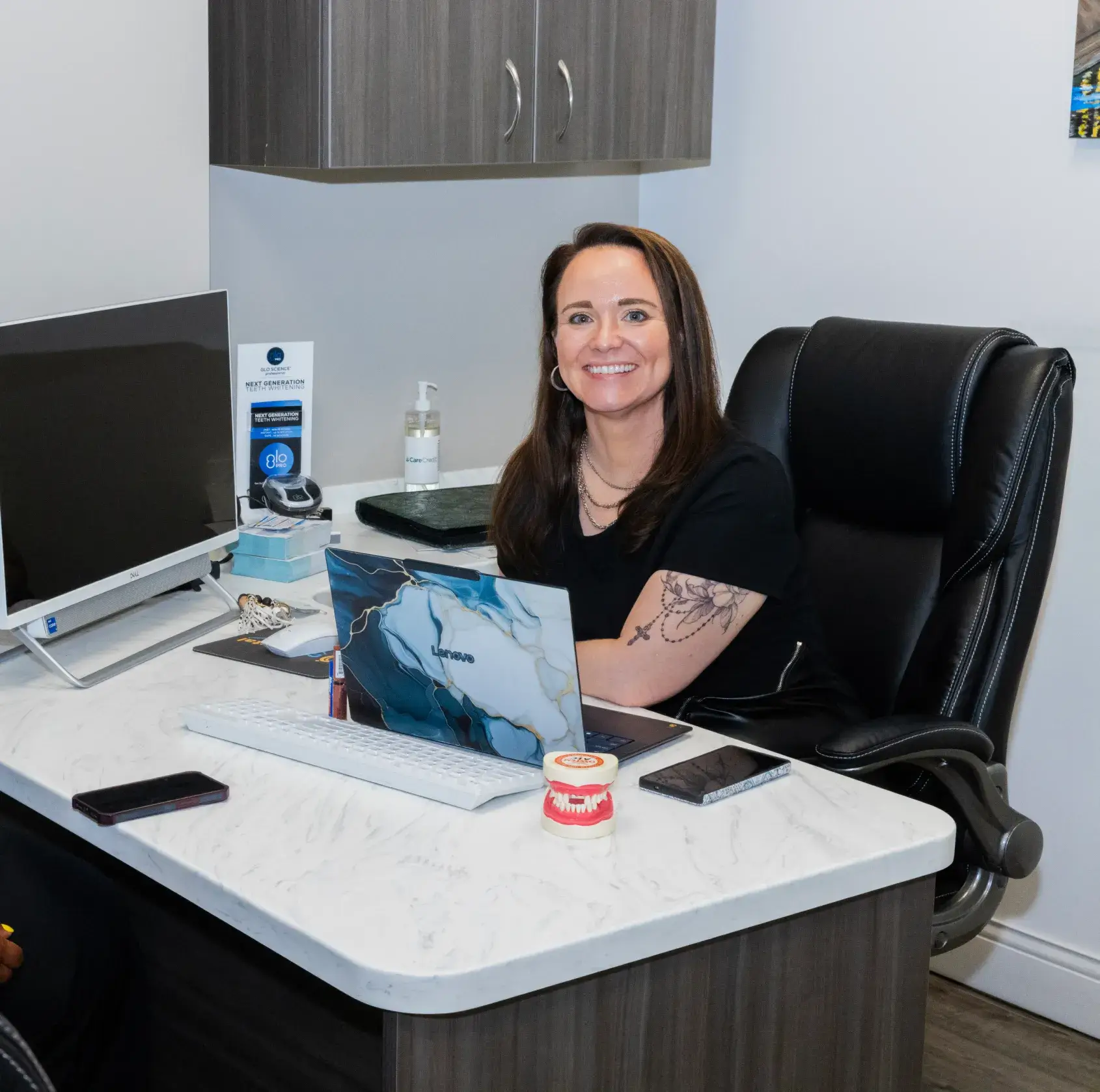 A woman sits smiling at a desk with a laptop and a computer monitor.