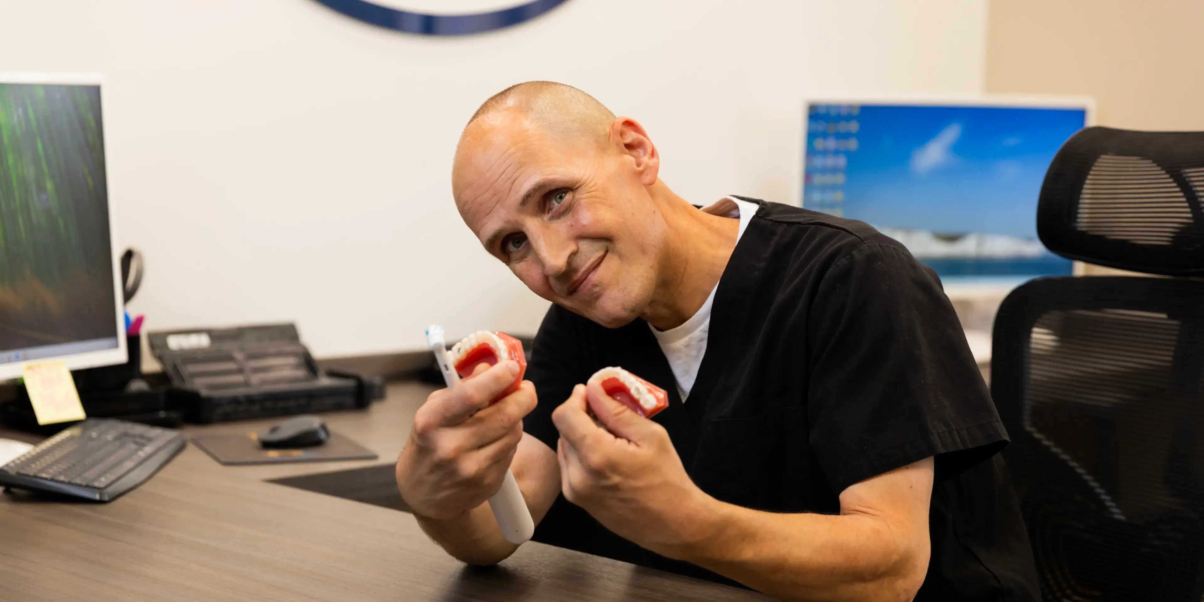 A person in scrubs, smiling and holding dental molds, sits at a desk in an office.