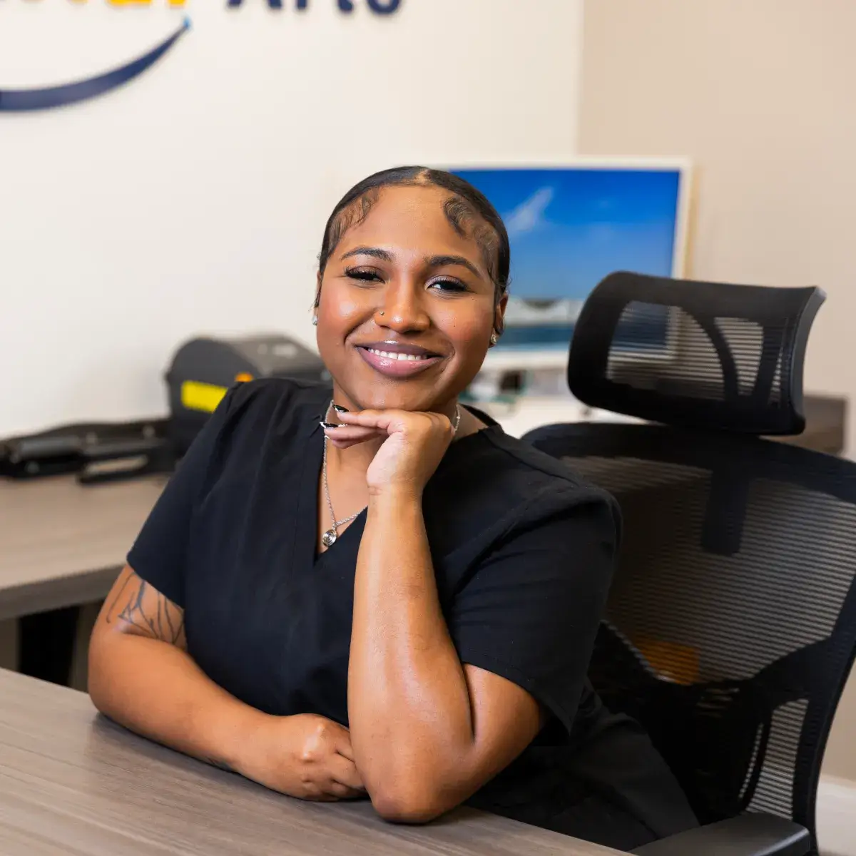 A person sitting at a desk, smiling, with a computer and office supplies in the background.