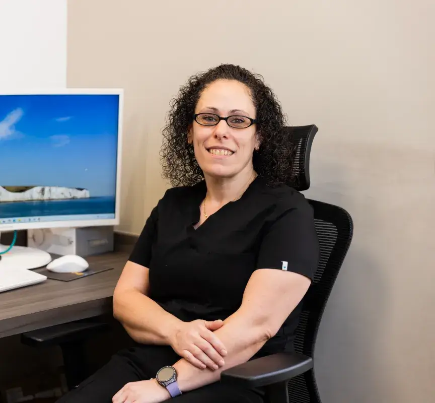 A person with curly hair and glasses sits smiling at a desk near a computer monitor.
