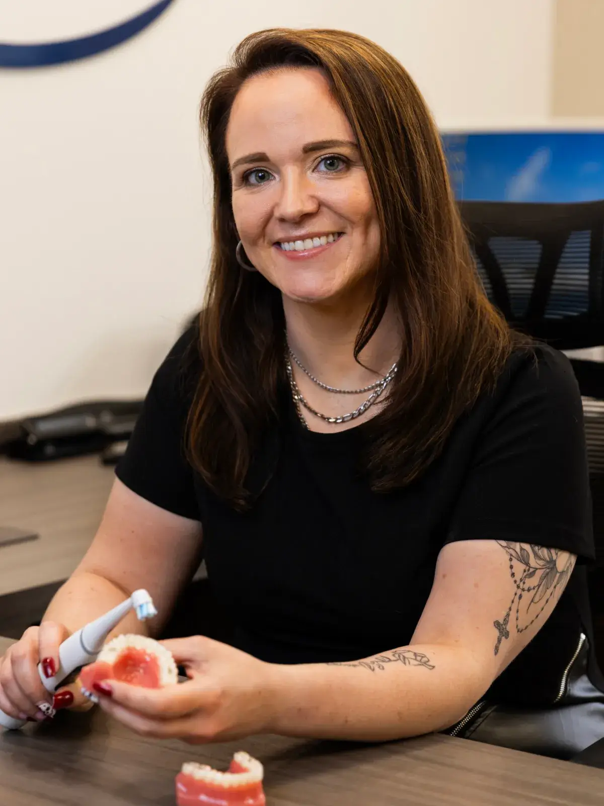 A woman smiles while holding a dental model and an electric toothbrush at a table.