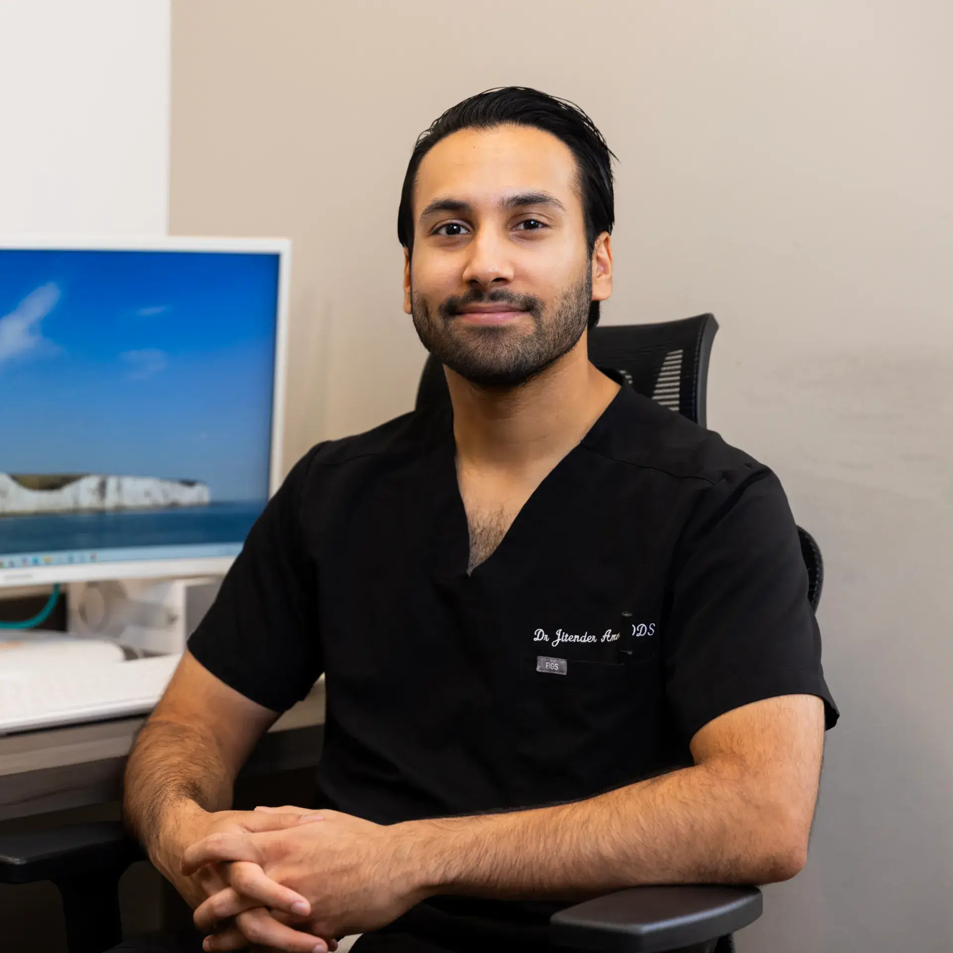 A man in black scrubs sits at a desk, facing the camera with a slight smile.