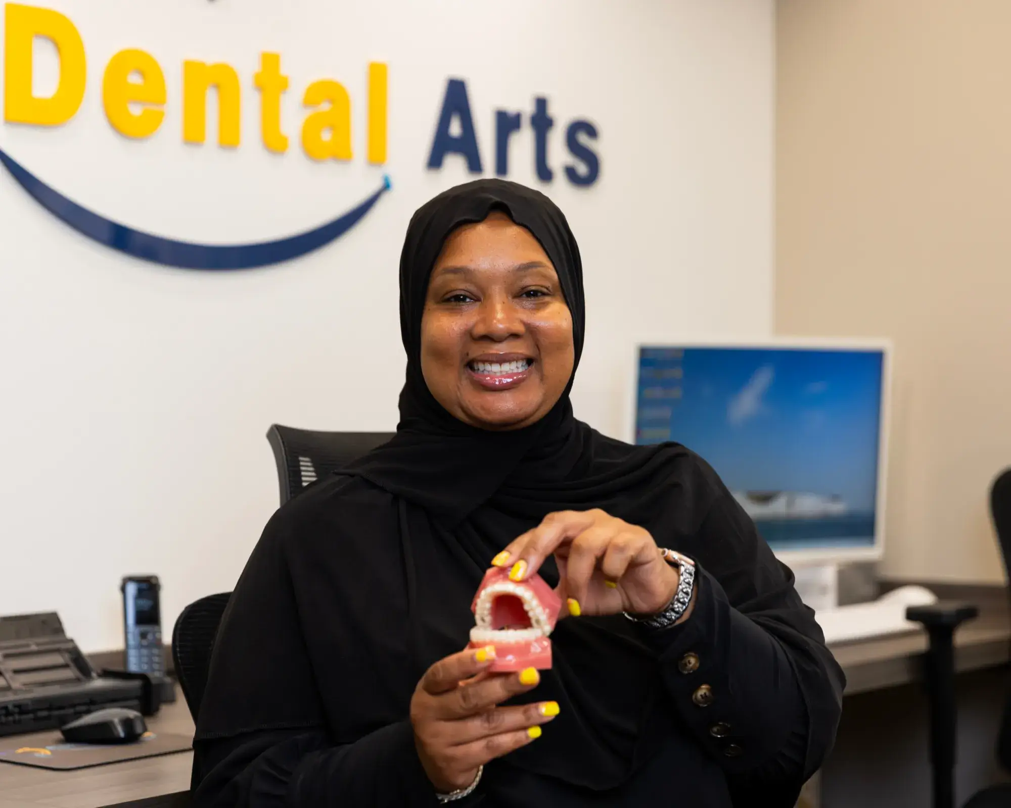 A smiling woman in a dental office holds a model of teeth, wearing a black outfit.