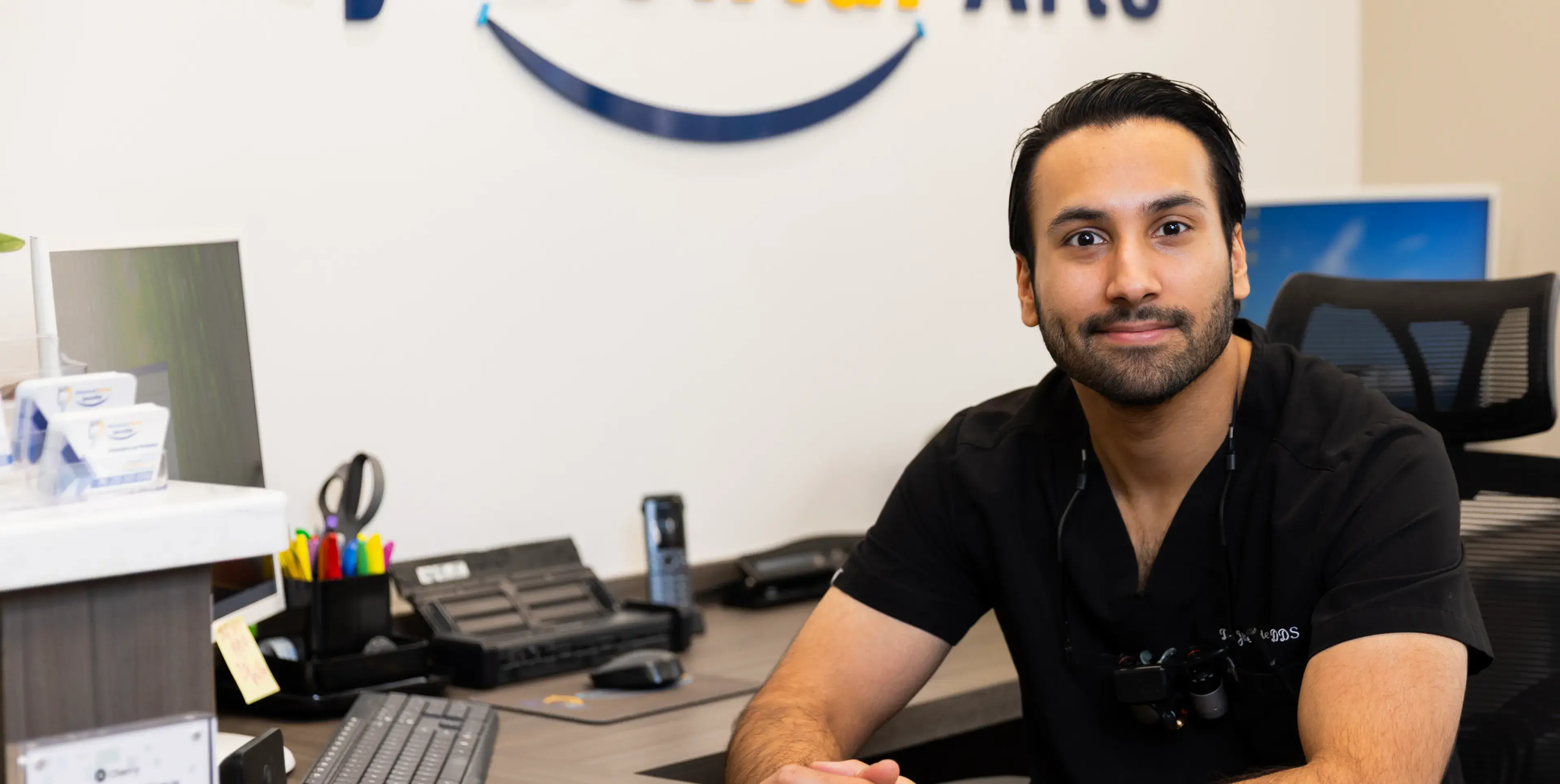 A man in a black shirt sits at a desk in an office, looking at the camera.
