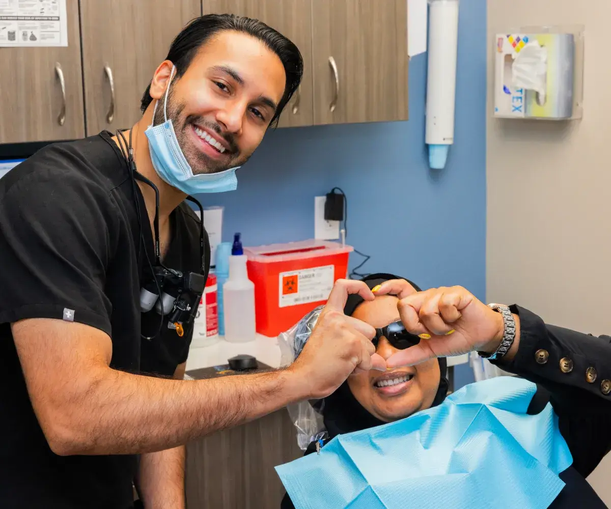 A dentist smiles while a patient in the chair forms a heart shape with their fingers.
